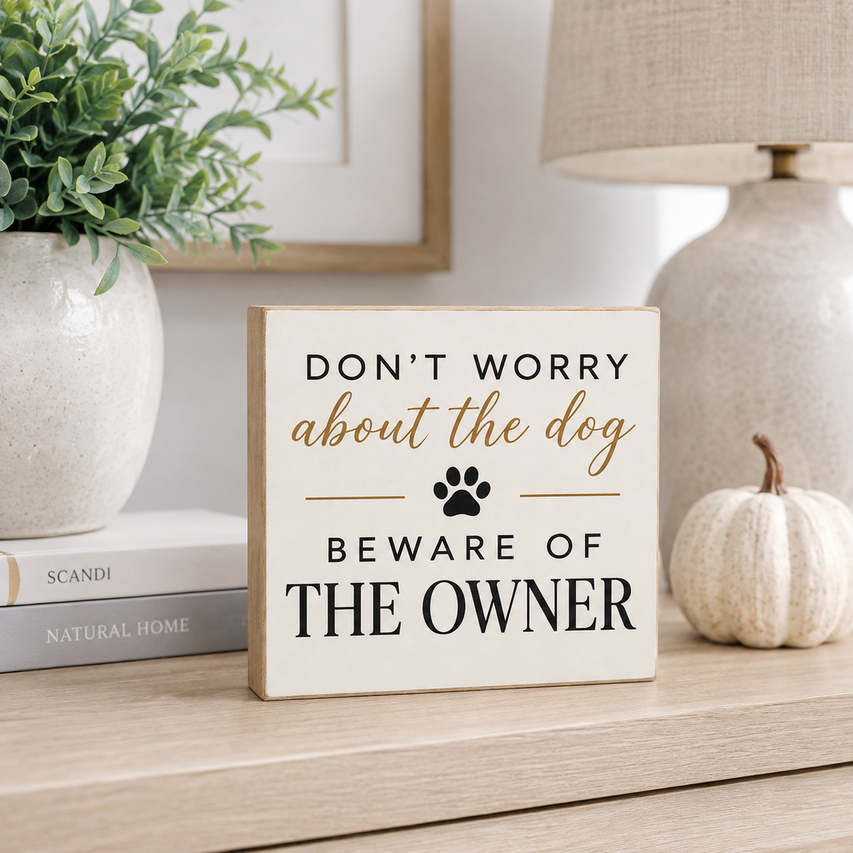 A square decorative sign with a light wood border and cream face reading “DON'T WORRY about the dog BEWARE OF THE OWNER” with a black paw print, displayed on a wooden shelf next to a small pumpkin, books, and a lamp.