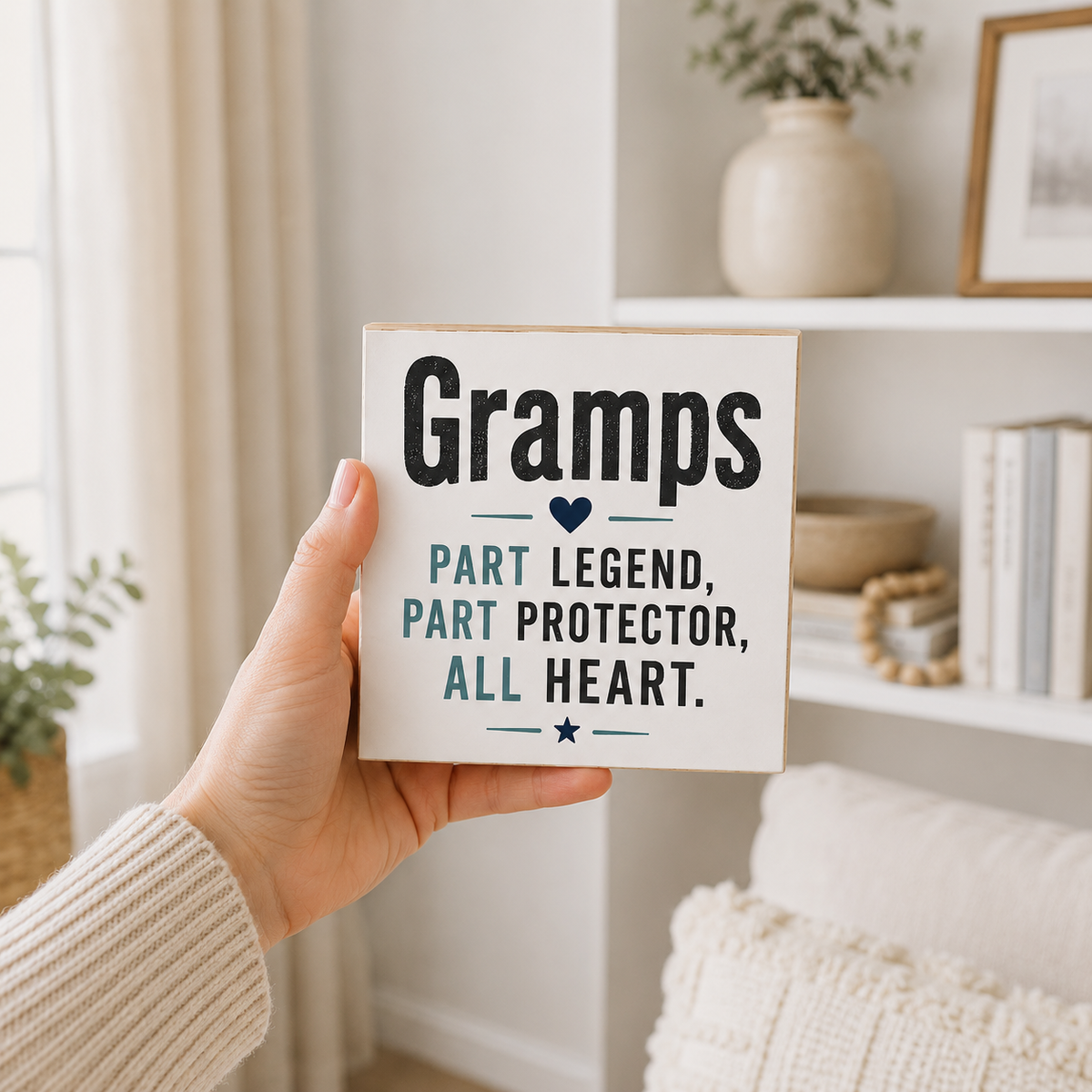 A hand holds a square white wooden sign with the text 'Gramps Part Legend, Part Protector, All Heart.' in black and navy lettering, set against a cozy, softly lit home interior background.