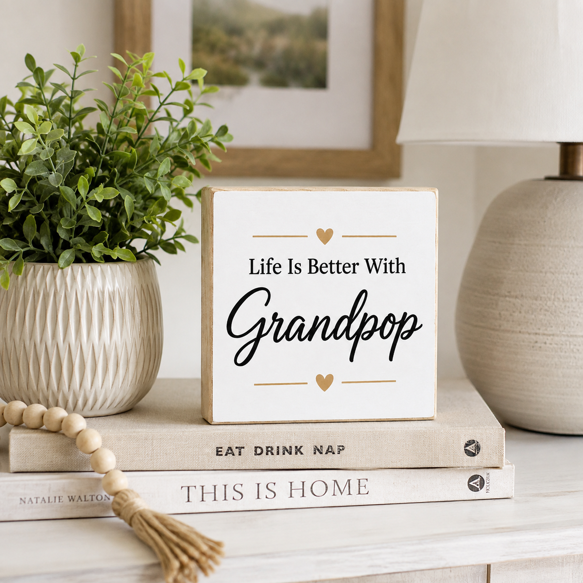 A square white decorative sign with natural wood edges and the text 'Life Is Better With Grandpop' in black script and serif lettering, styled on stacked books beside a potted plant and a lamp.