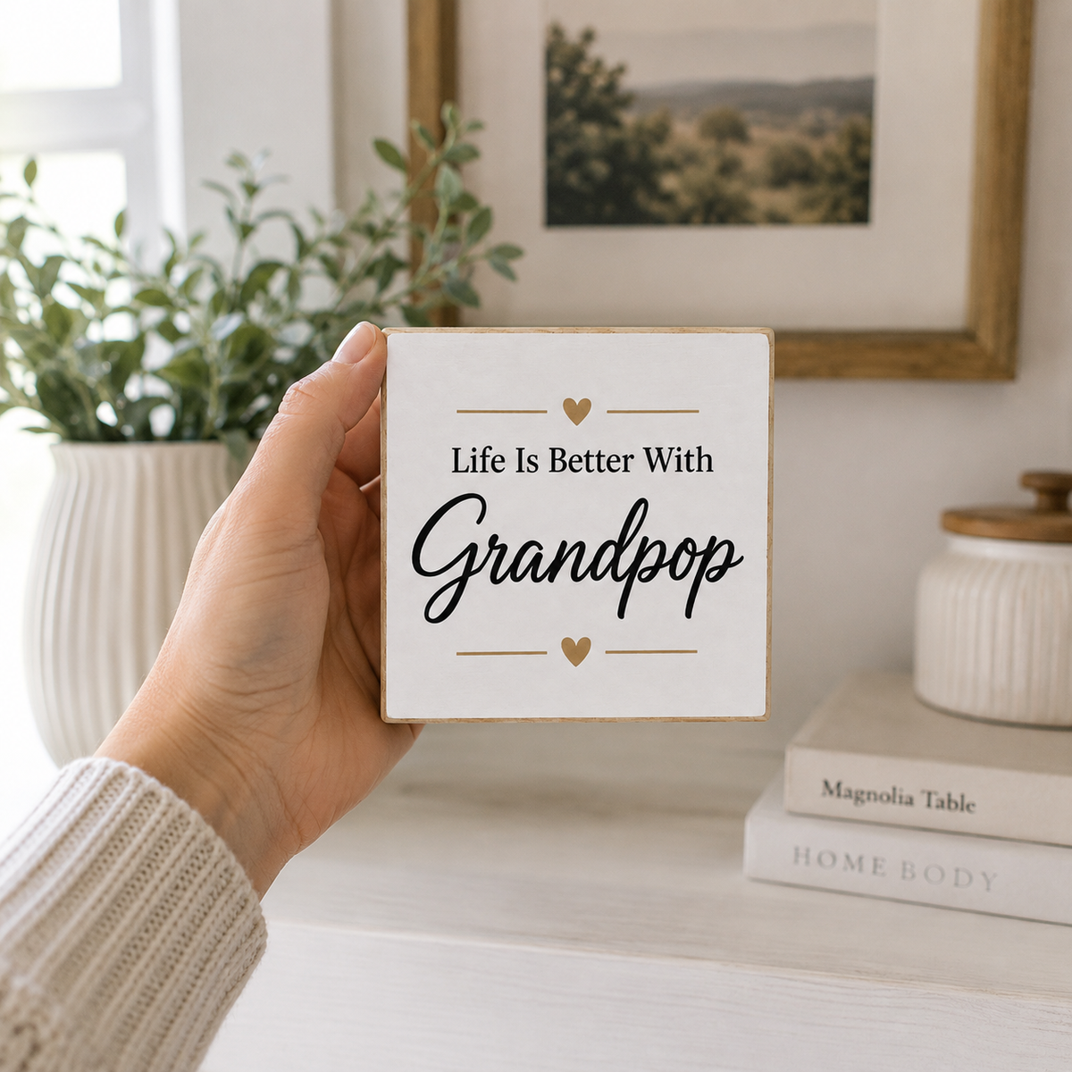 A hand holds a square white decorative wooden sign with the text 'Life Is Better With Grandpop' in black script and gold heart accents, set against a softly blurred home interior background with a plant, framed picture, and books.