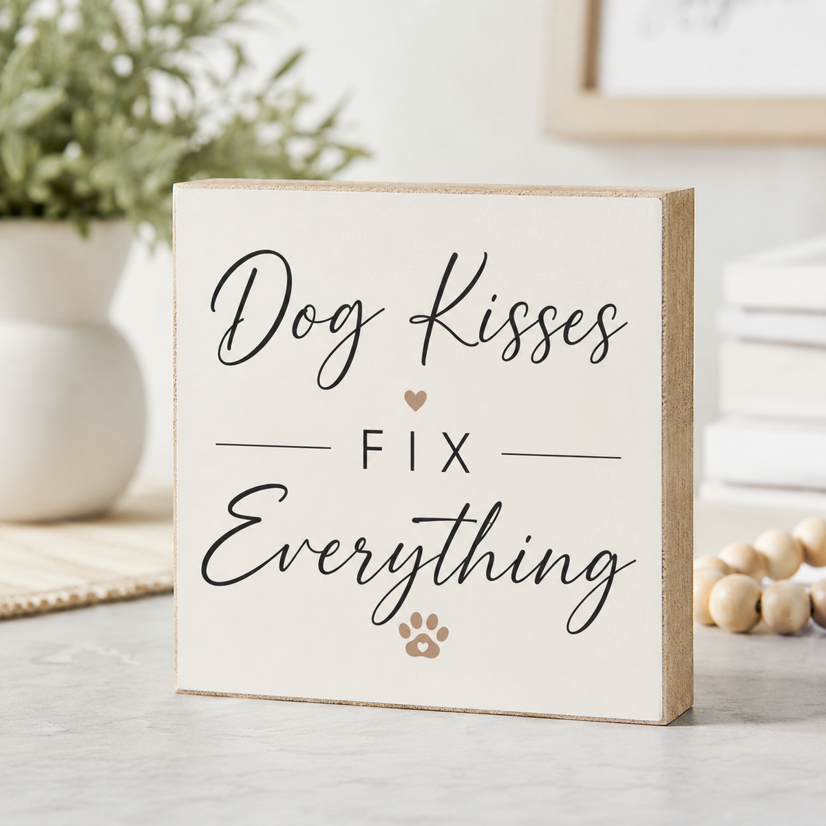 A square decorative wooden sign with a natural wood edge and cream face, printed with the phrase 'Dog Kisses FIX Everything' in black script and serif lettering, plus a small heart and paw print. The sign is photographed on a tabletop with a blurred plant, books, and beads in the background.