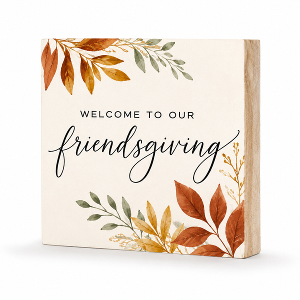 Square wooden sign with a cream face and natural wood side edges, printed with the phrase “WELCOME TO OUR friendsgiving” in black text, surrounded by autumn leaves in orange, brown, and muted green.