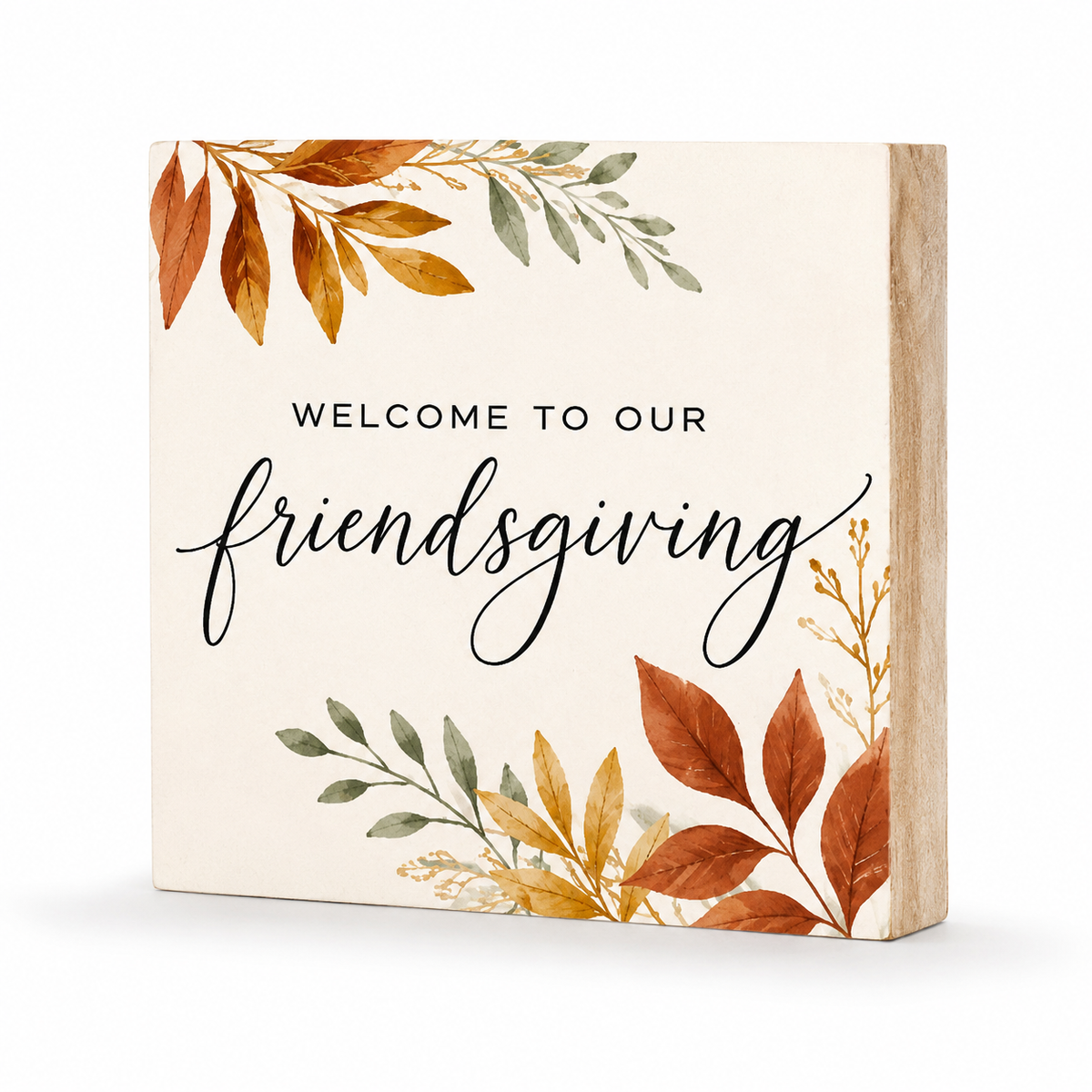 Square wooden sign with a cream face and natural wood side edges, printed with the phrase “WELCOME TO OUR friendsgiving” in black text, surrounded by autumn leaves in orange, brown, and muted green.