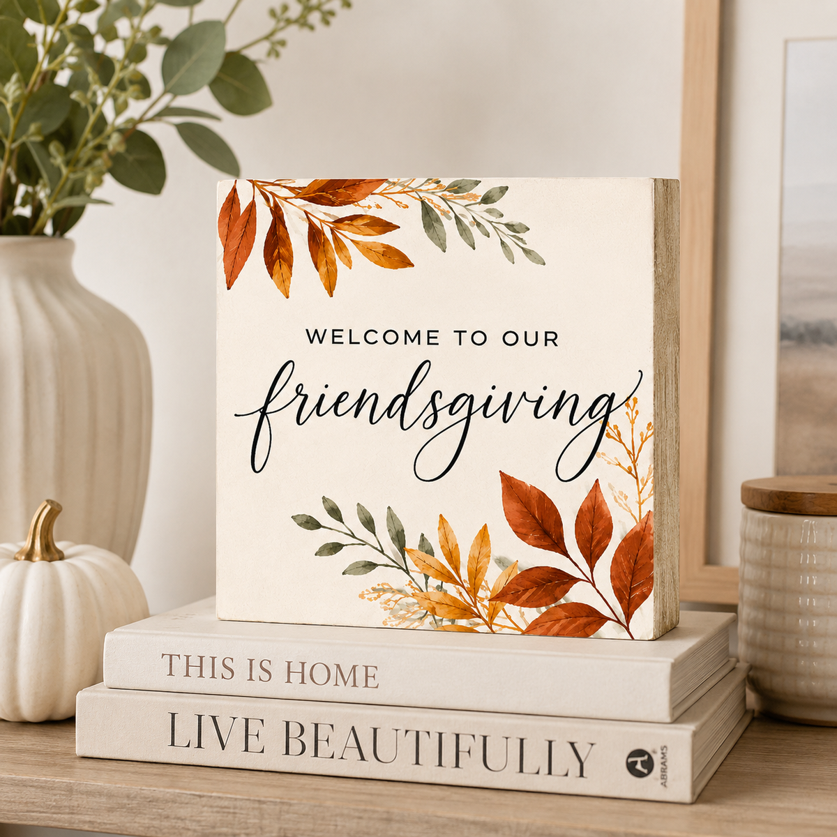 A square decorative sign with the words “WELCOME TO OUR friendsgiving” in black script and serif lettering, decorated with watercolor-style autumn leaves in orange, gold, and green on an off-white background. The sign is styled on top of two books on a wooden shelf, with a ceramic vase, greenery, and a small white pumpkin in the background.