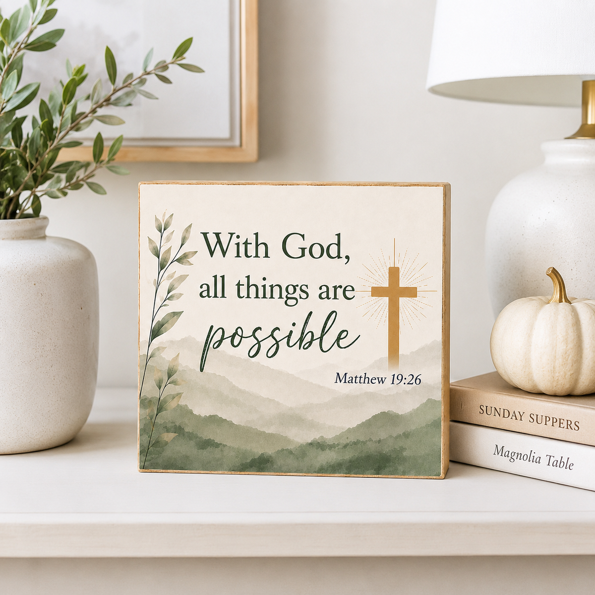 A square decorative wall sign with the phrase 'With God, all things are possible Matthew 19:26' and a wooden cross, styled on a shelf beside a vase, framed art, a lamp, books, and a small pumpkin.