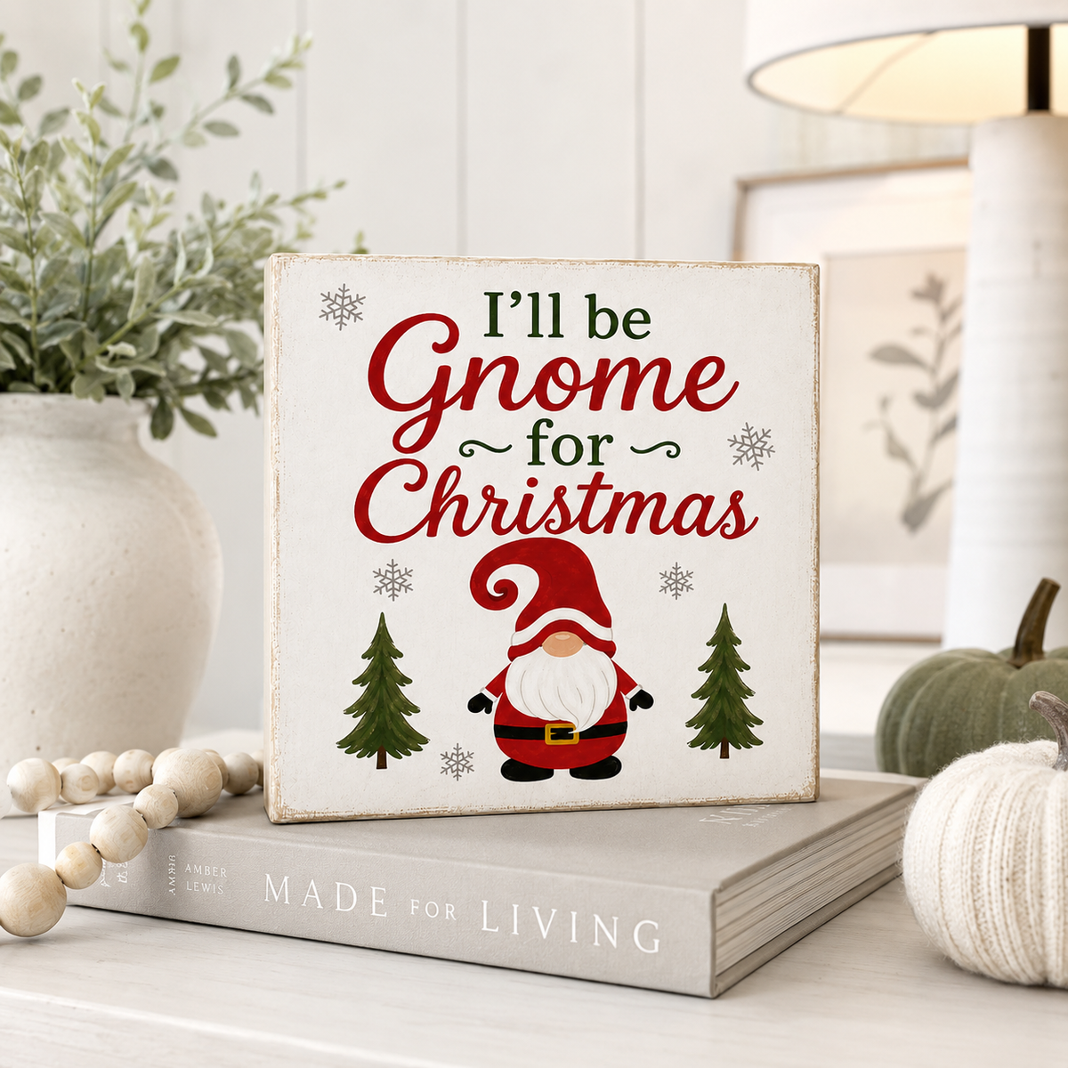 A square decorative sign with the phrase “I’ll be Gnome for Christmas” in red and green lettering, featuring a small gnome illustration and trees, displayed on a book in a styled holiday interior setting.