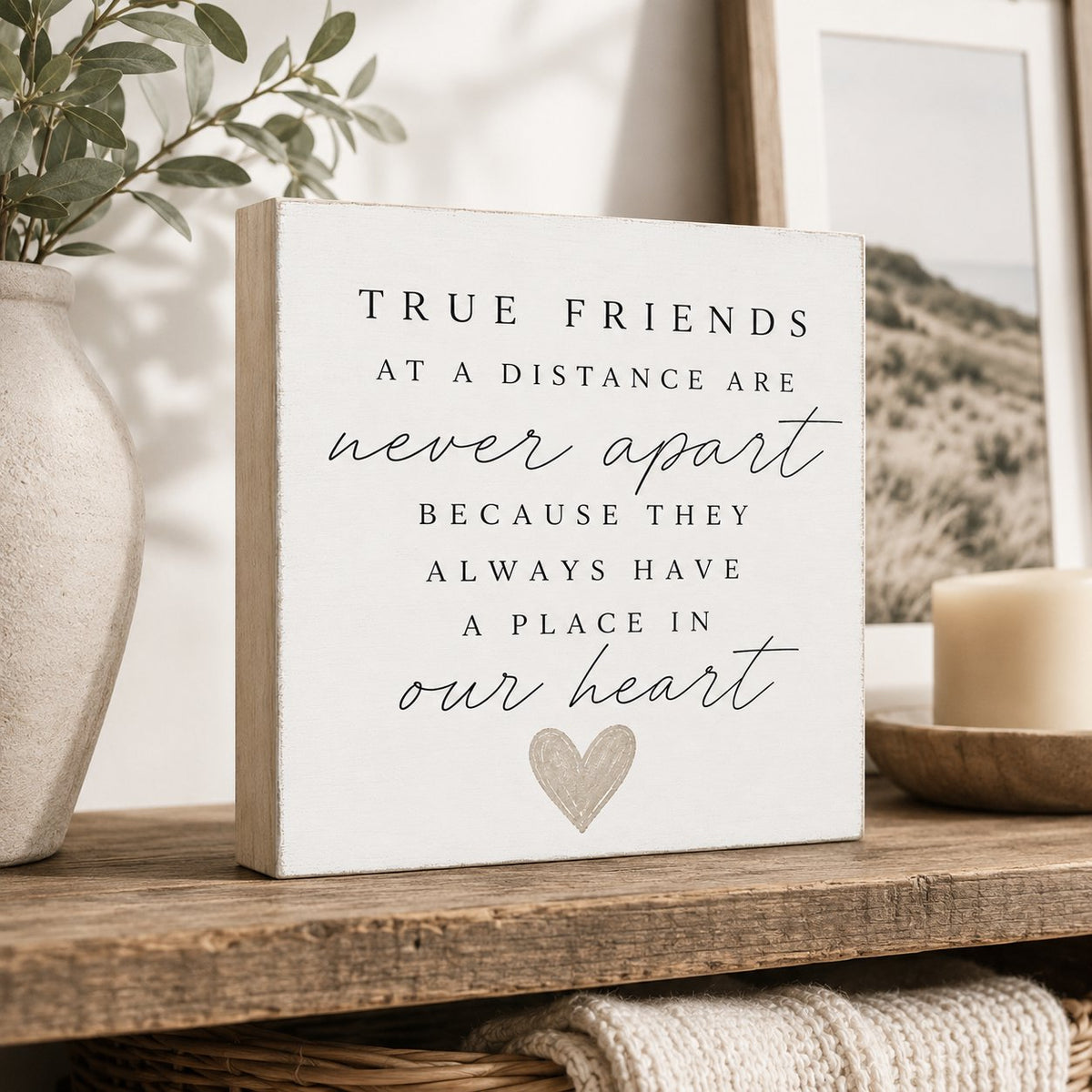 A square white decorative sign with a light wood edge reads 'TRUE FRIENDS AT A DISTANCE ARE never apart BECAUSE THEY ALWAYS HAVE A PLACE IN our heart' above a small heart illustration. The sign is styled on a wooden shelf with a vase, greenery, and a candle in a softly lit interior setting.