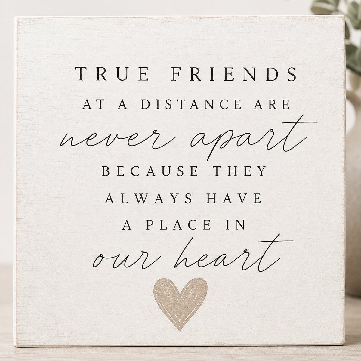 A square white wooden sign with black printed text reading 'TRUE FRIENDS AT A DISTANCE ARE never apart BECAUSE THEY ALWAYS HAVE A PLACE IN our heart' and a small glittery heart at the bottom, shown against a softly blurred indoor background.