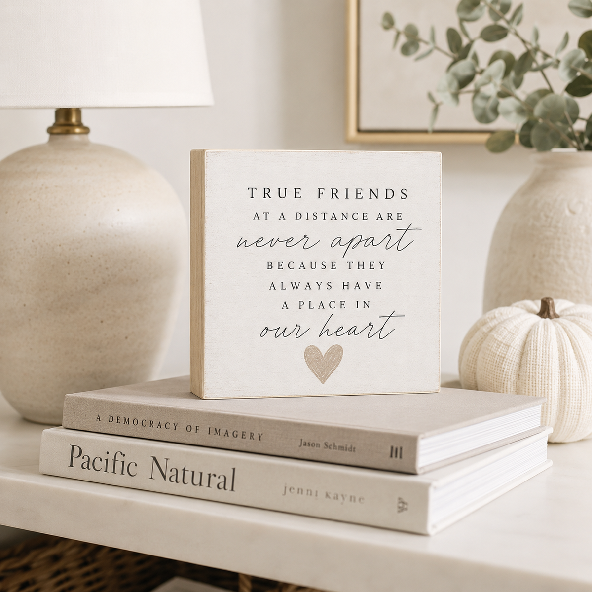 A square cream-colored decorative sign with black text reading 'TRUE FRIENDS AT A DISTANCE ARE never apart BECAUSE THEY ALWAYS HAVE A PLACE IN our heart' and a small heart illustration, styled on top of two books on a shelf beside a lamp and a small pumpkin.