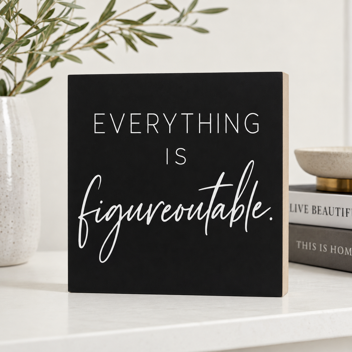 A square black decorative sign with the phrase 'EVERYTHING IS figureoutable.' in white lettering, styled on a white tabletop with a vase and books in the background.