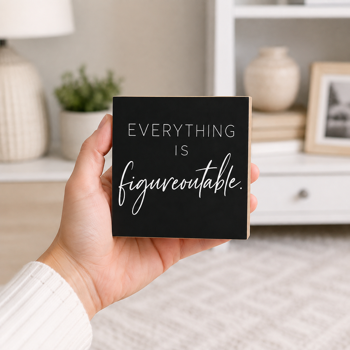 A hand holds a square black sign with the phrase 'EVERYTHING IS figureoutable.' in white lettering, set against a softly blurred home interior background.