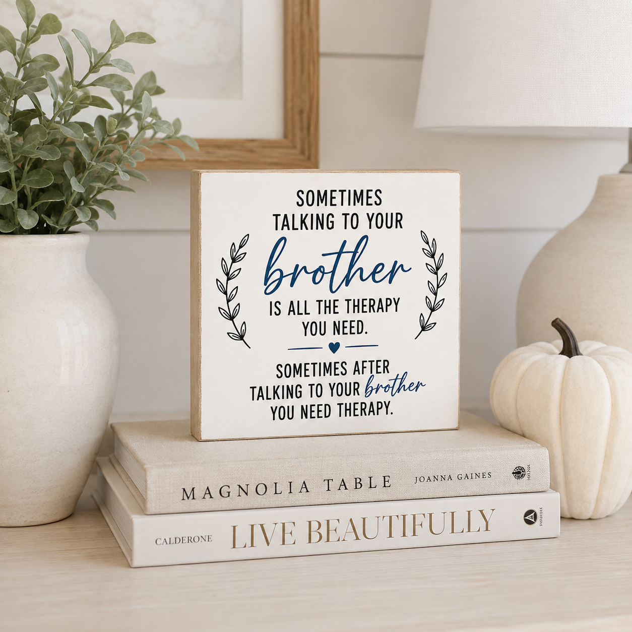 A square wooden sign with a humorous quote for brothers, styled on a shelf next to a potted plant and a small pumpkin.