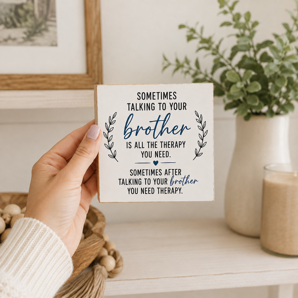 A hand holds a square white decorative sign with navy text that reads, 'Sometimes talking to your brother is all the therapy you need. Sometimes after talking to your brother you need therapy.