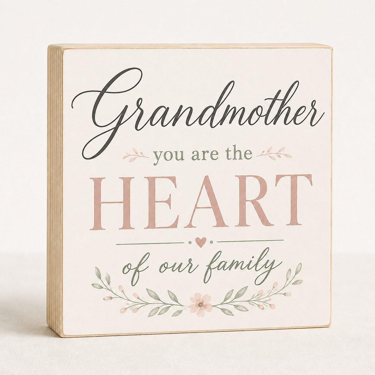 A square decorative wooden sign with a cream face and natural wood edges, printed with the phrase 'Grandmother you are the HEART of our family' in black and dusty pink lettering, with small floral accents, on a light neutral background.