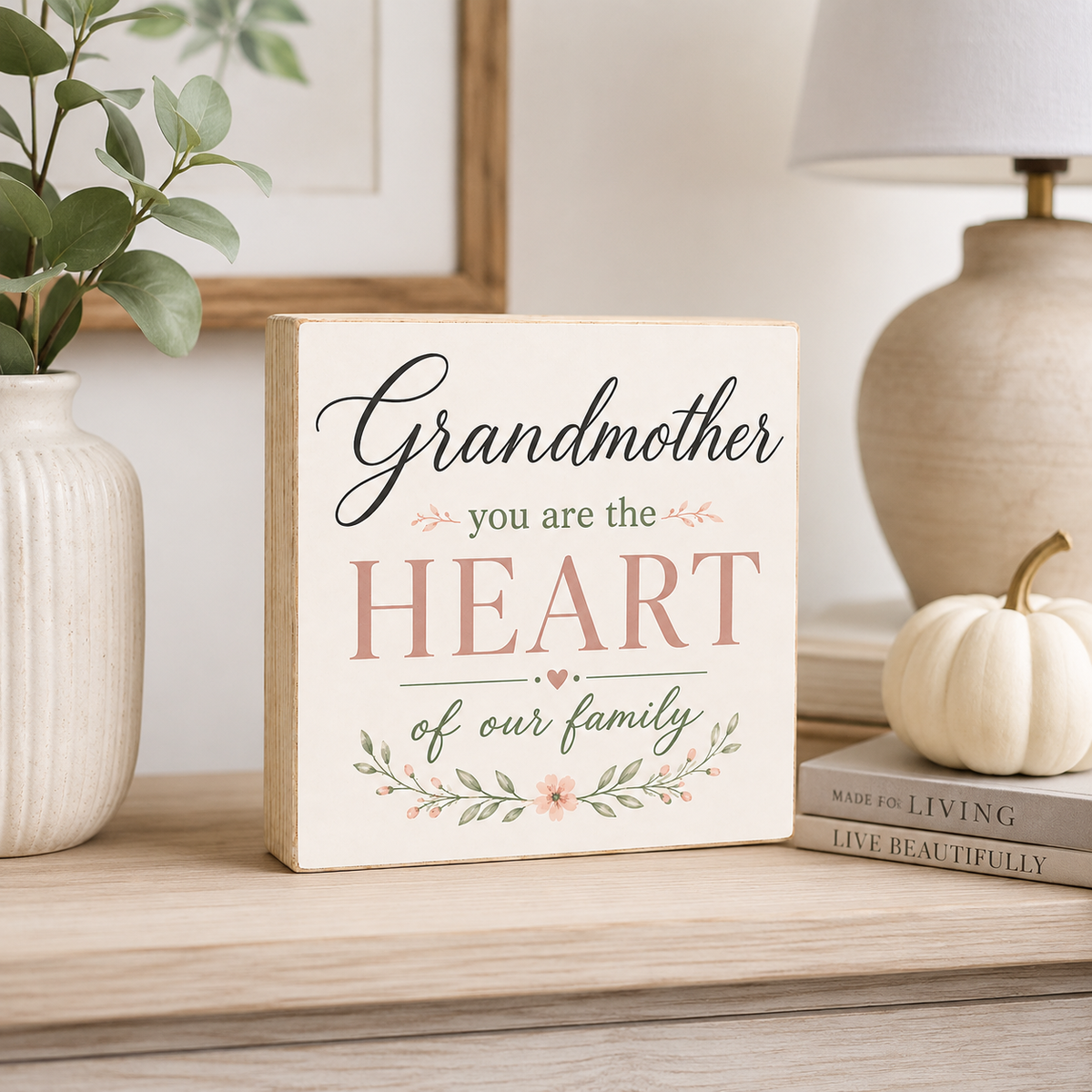 A rectangular wooden sign with the phrase 'Grandmother you are the HEART of our family' in black and dusty pink lettering, decorated with a small floral vine, displayed on a wooden shelf next to a vase, framed art, a lamp, and a small pumpkin.