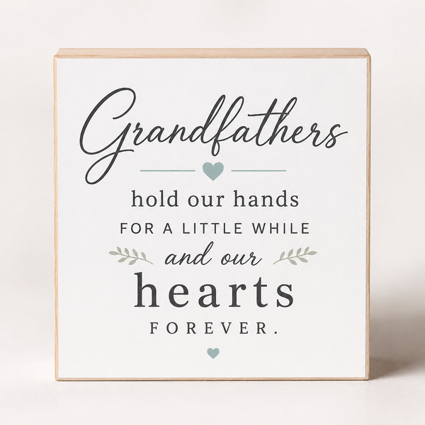 A square wooden sign with a white face and natural wood edges, featuring the printed message 'Grandfathers hold our hands for a little while and our hearts FOREVER' in dark gray lettering with small sage green heart and leaf accents, shown on a light neutral background.