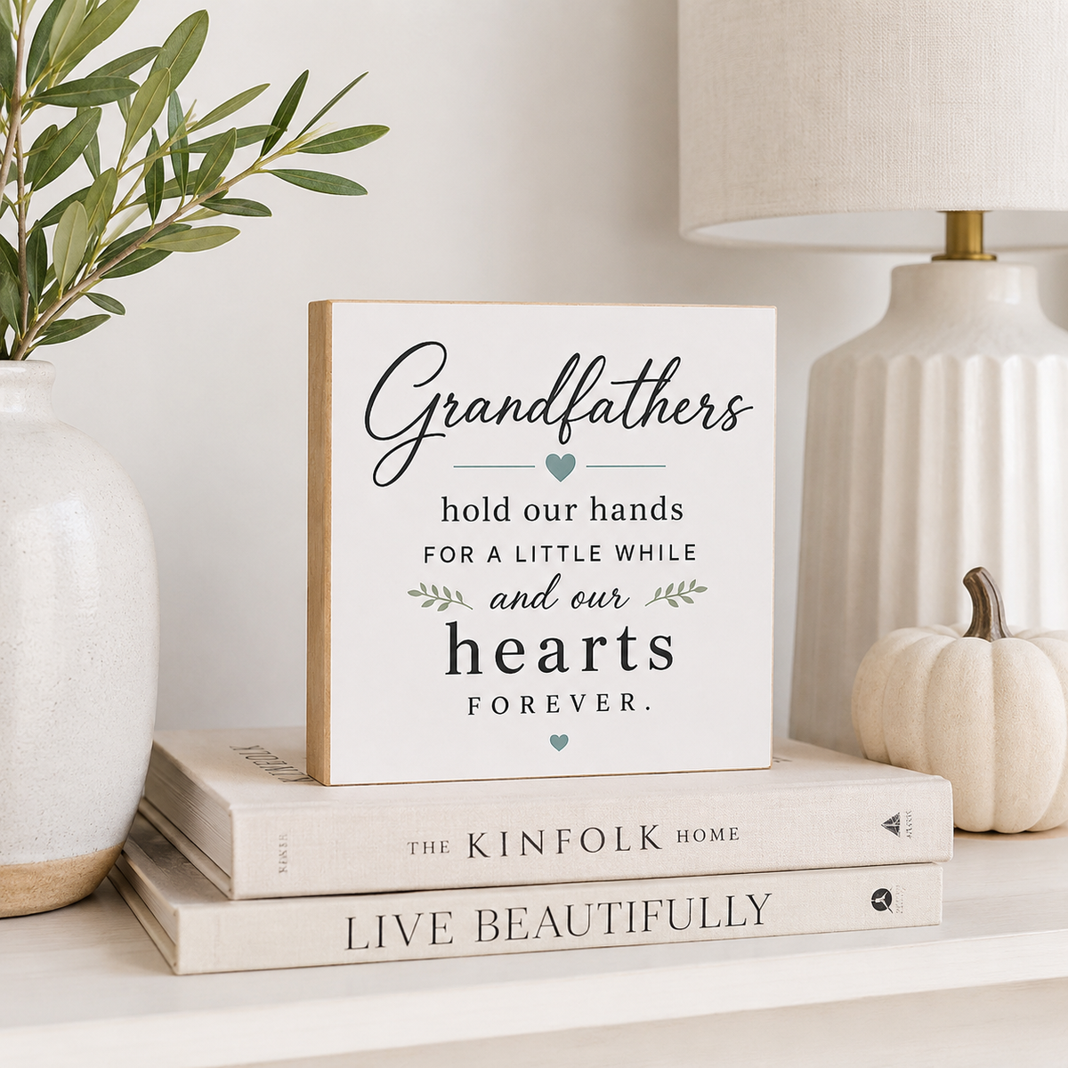A square white decorative sign with a light wood edge and black script text reading 'Grandfathers hold our hands for a little while and our hearts FOREVER' with small teal heart and leaf accents, styled on stacked books beside a vase with greenery and a lamp.