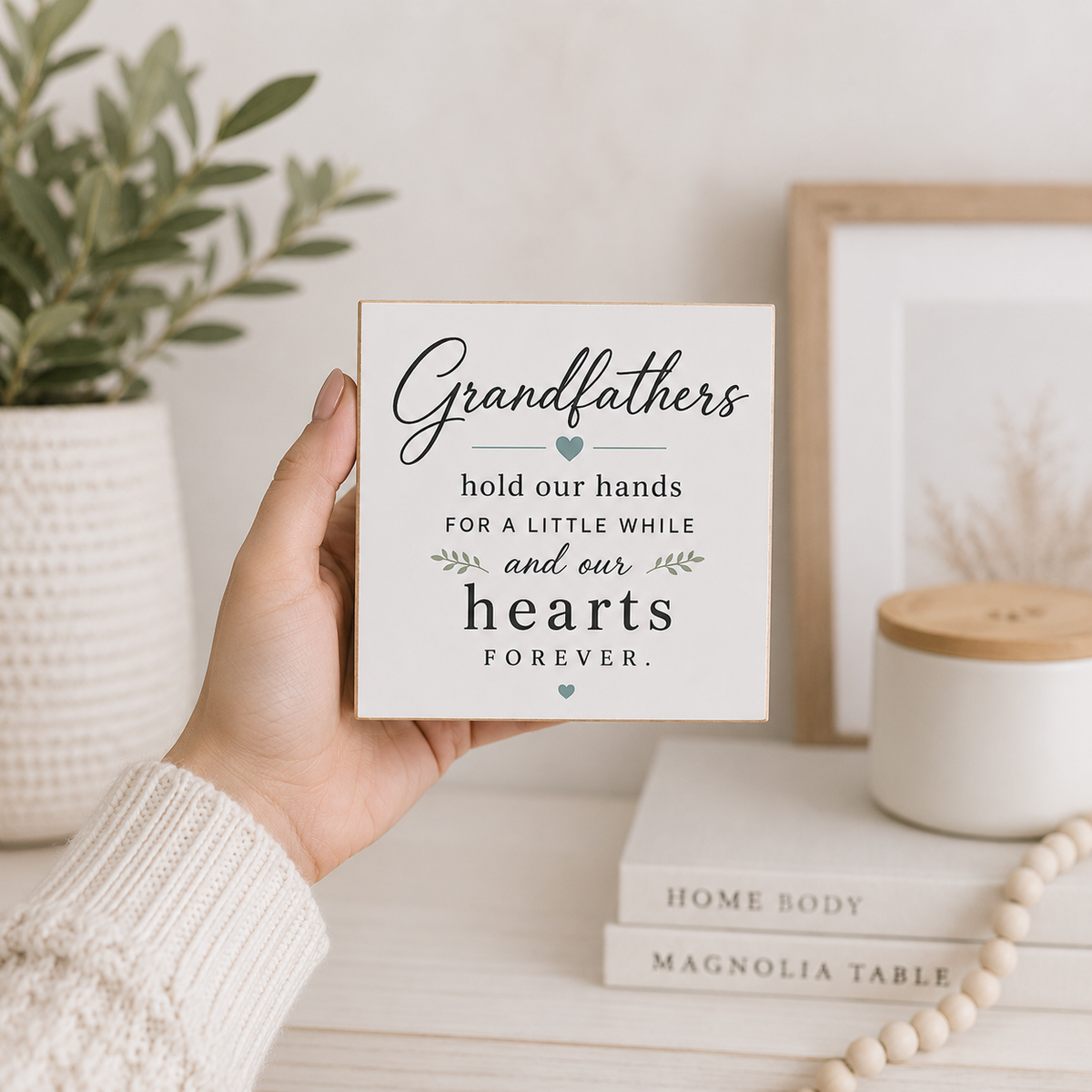 A hand in a cream knit sweater holds a square white decorative sign with a warm wood-toned edge. The sign reads, 'Grandfathers hold our hands for a little while and our hearts FOREVER.