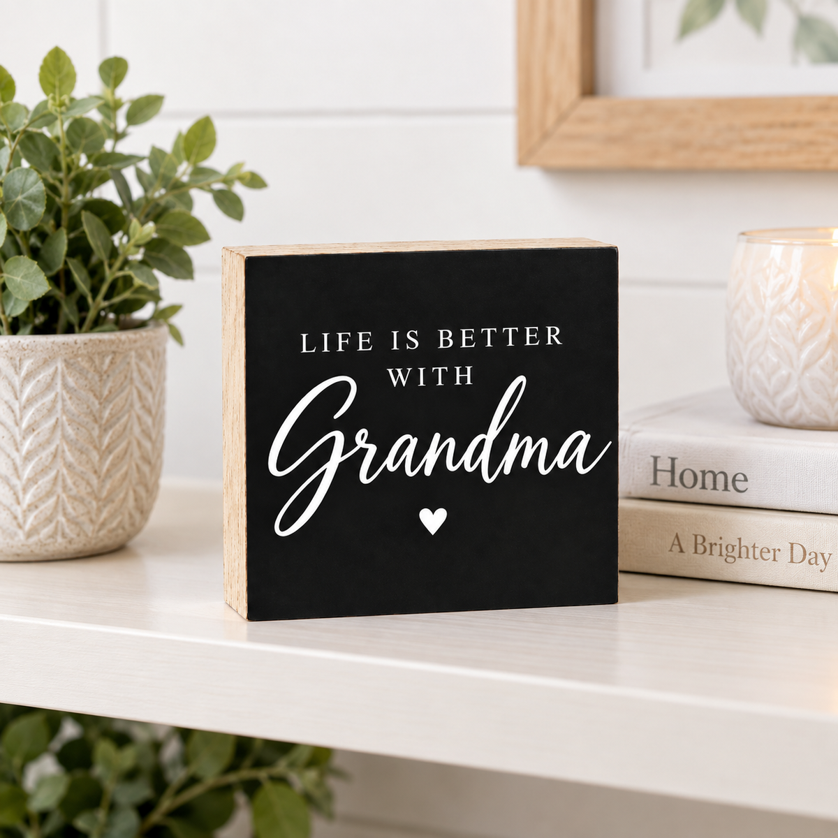 A square black wooden sign with white lettering that reads 'LIFE IS BETTER WITH Grandma' and a small heart, displayed on a white shelf with a potted plant, books, and a candle in a home setting.