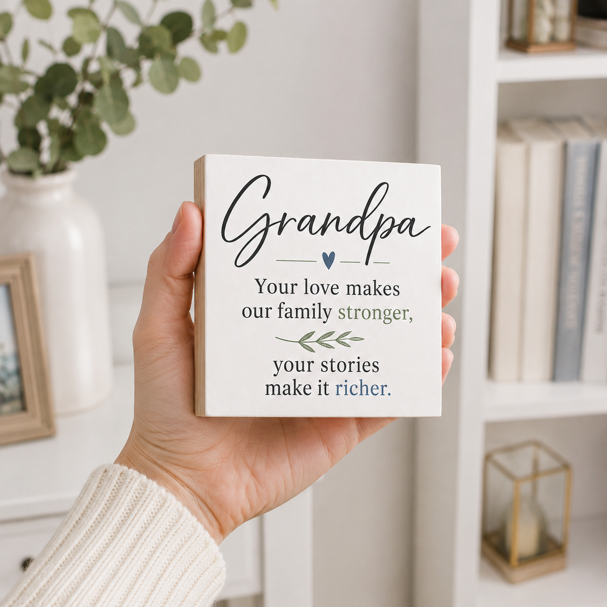 A hand holding a square white wooden plaque with a personalized quote for 'Grandpa' in black, green, and blue text, set against a cozy, softly blurred home interior background.