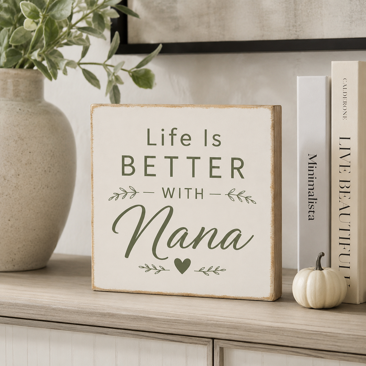 A square decorative sign with the phrase “Life Is BETTER WITH Nana” in dark green lettering on a warm off-white background, styled on a light wood shelf beside a vase, books, and a small pumpkin.