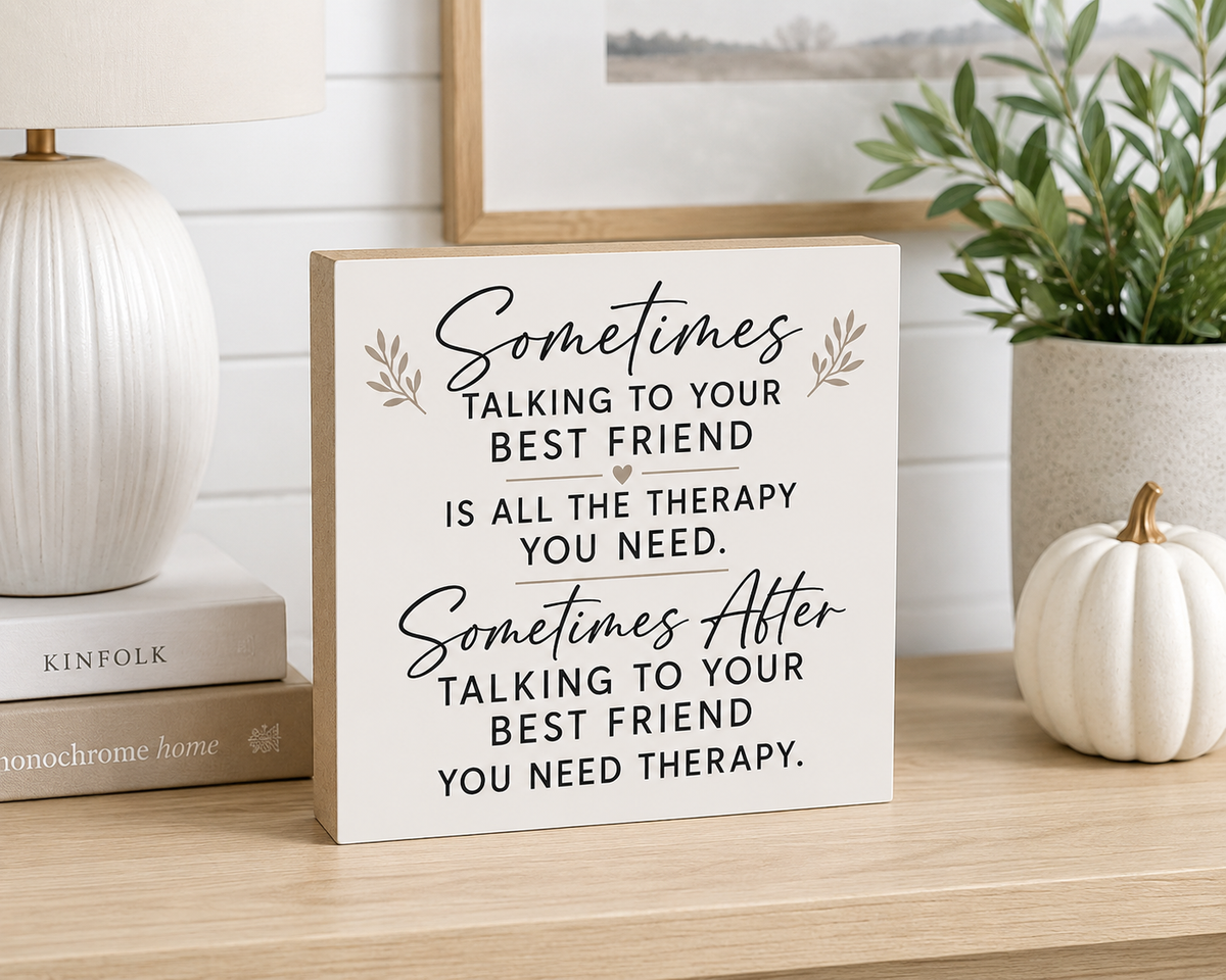 A square wooden sign with a warm off-white finish displays a therapy-themed quote in black script and serif text, styled on a wooden shelf next to a white ceramic lamp, a small white pumpkin, and a potted green plant.