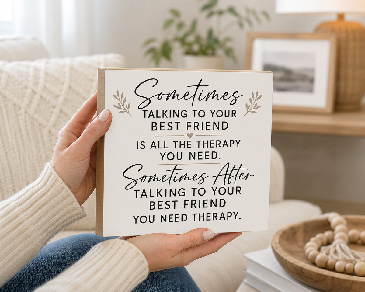 A person holds a square white sign with a beige wood-look border and black text that reads, "Sometimes Talking to Your Best Friend Is All the Therapy You Need. Sometimes After Talking to Your Best Friend You Need Therapy.
