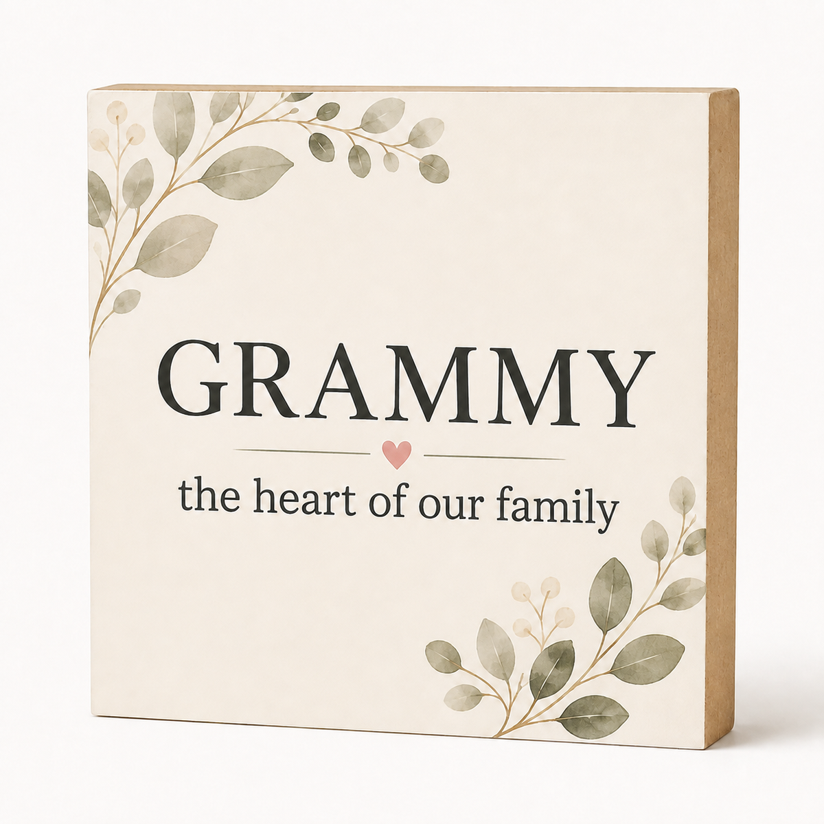 A square decorative sign with the words “GRAMMY the heart of our family” in black text on a warm cream background, decorated with soft green and gray botanical leaves in the corners and a small pink heart. The sign has a natural kraft wood side edge and is photographed on a clean white background.