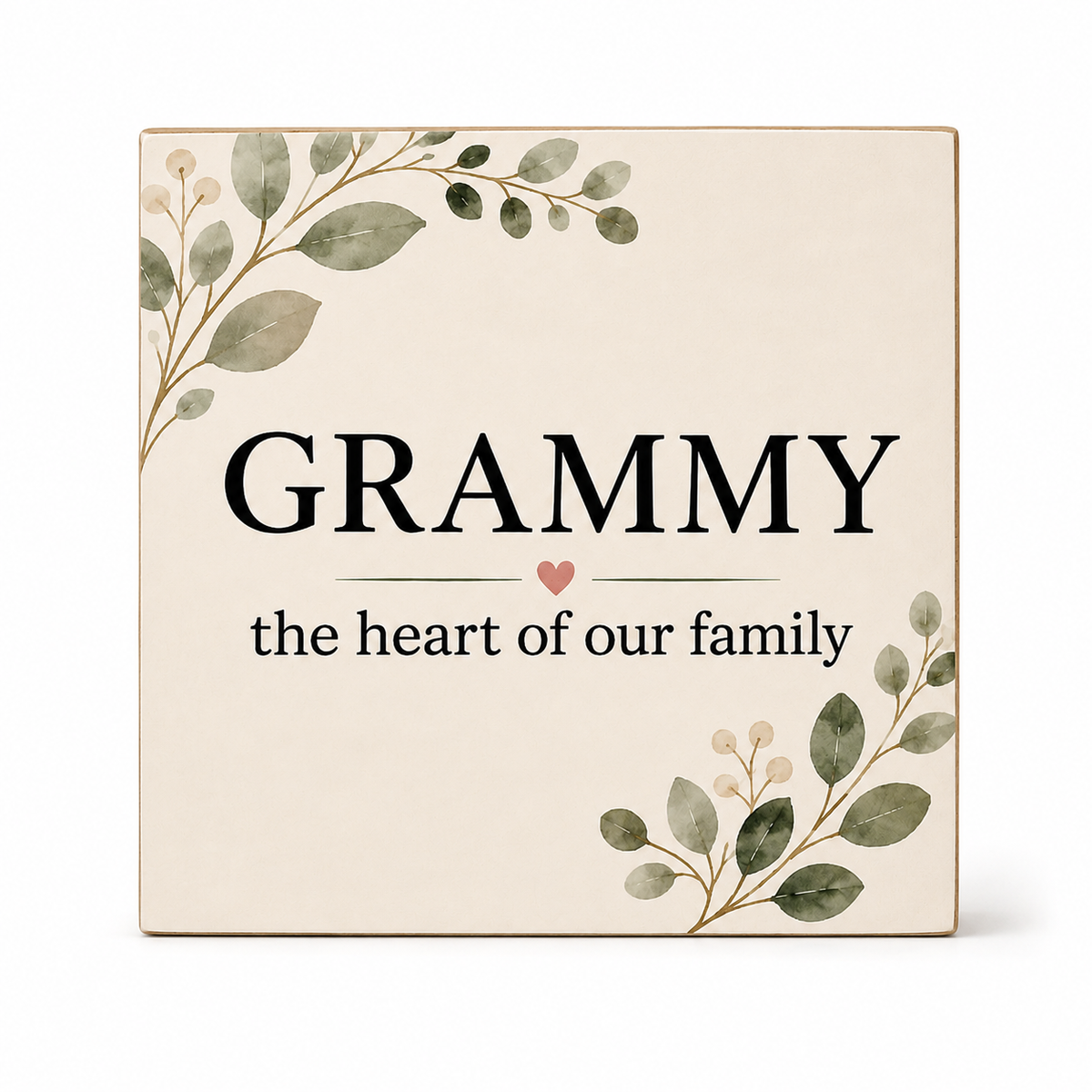 A square decorative plaque or sign with the word “GRAMMY” in large black letters and the phrase “the heart of our family” below, framed by soft green botanical illustrations in the top left and bottom right corners. The sign has a warm cream background and is photographed on a clean white background.