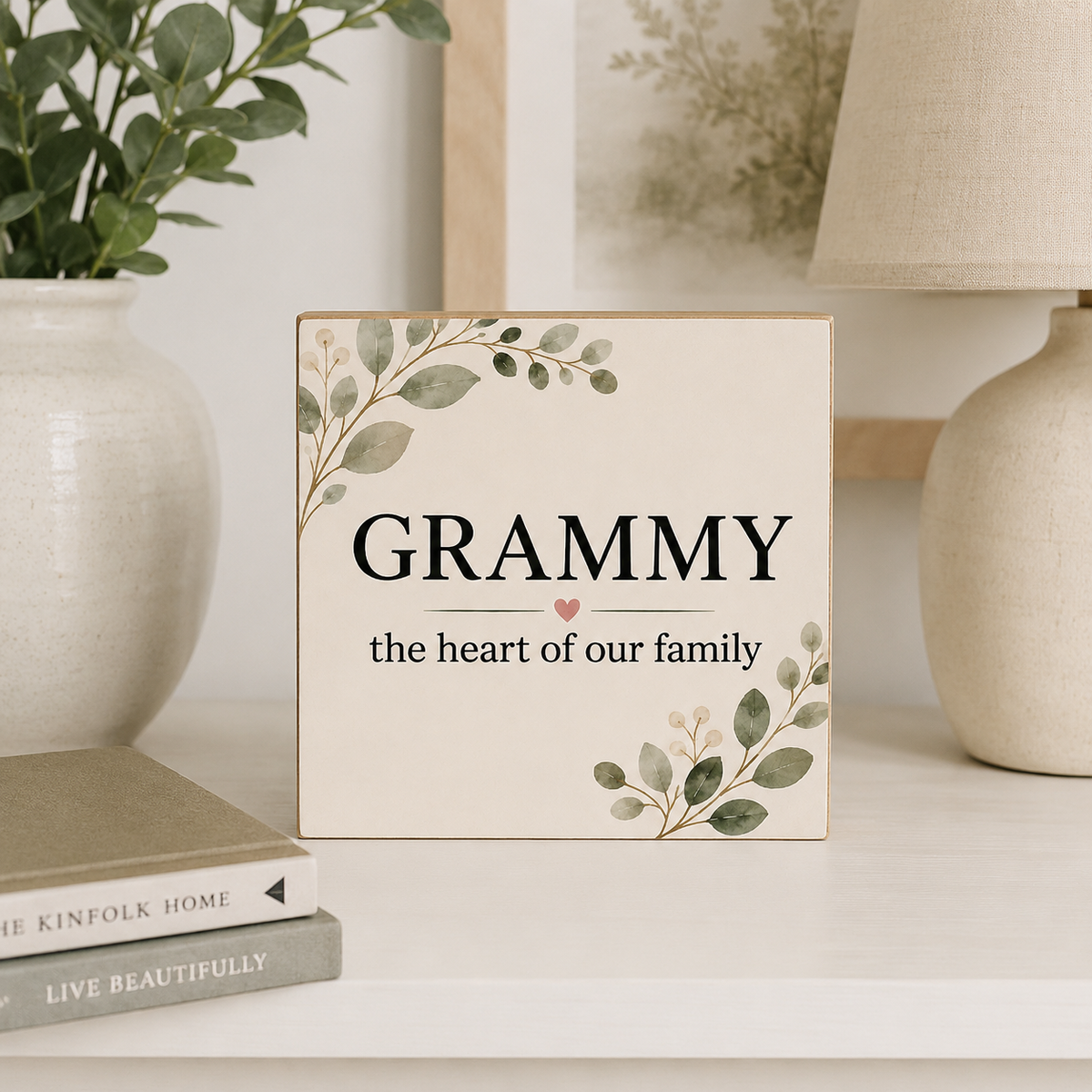 A square decorative sign with the word “GRAMMY” and the phrase “the heart of our family” in black text, decorated with green leaf illustrations in the corners, displayed on a tabletop in a styled home setting.