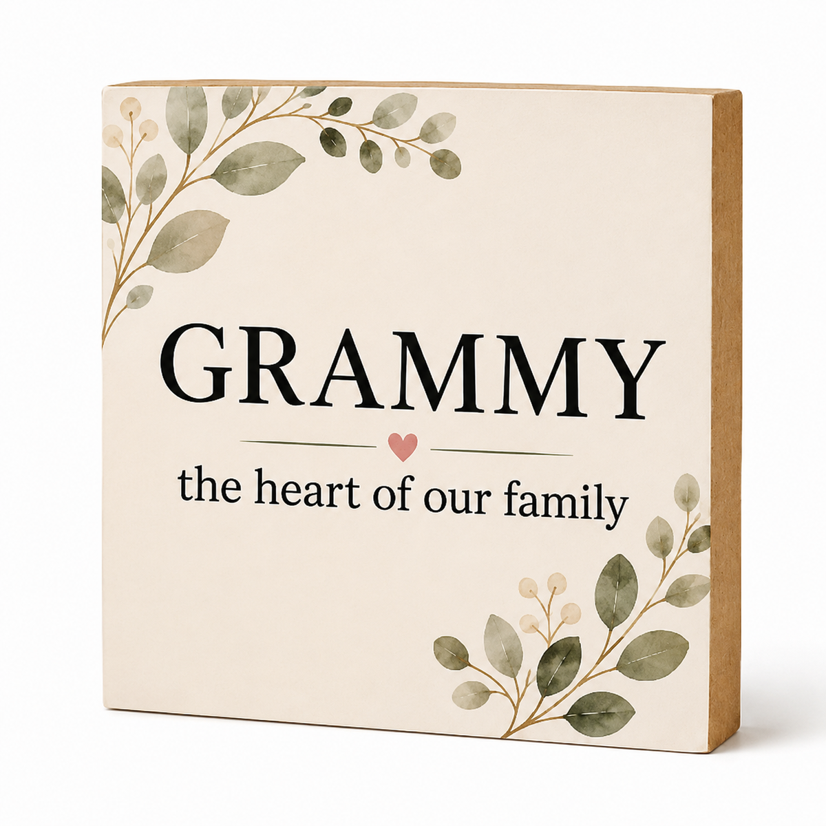 A square decorative sign with a cream face, printed with the word “GRAMMY” in large black letters and the phrase “the heart of our family” below, accented by a small pink heart and botanical leaf illustrations in muted green and beige. The sign has a natural wood side edge and is photographed on a clean white background.