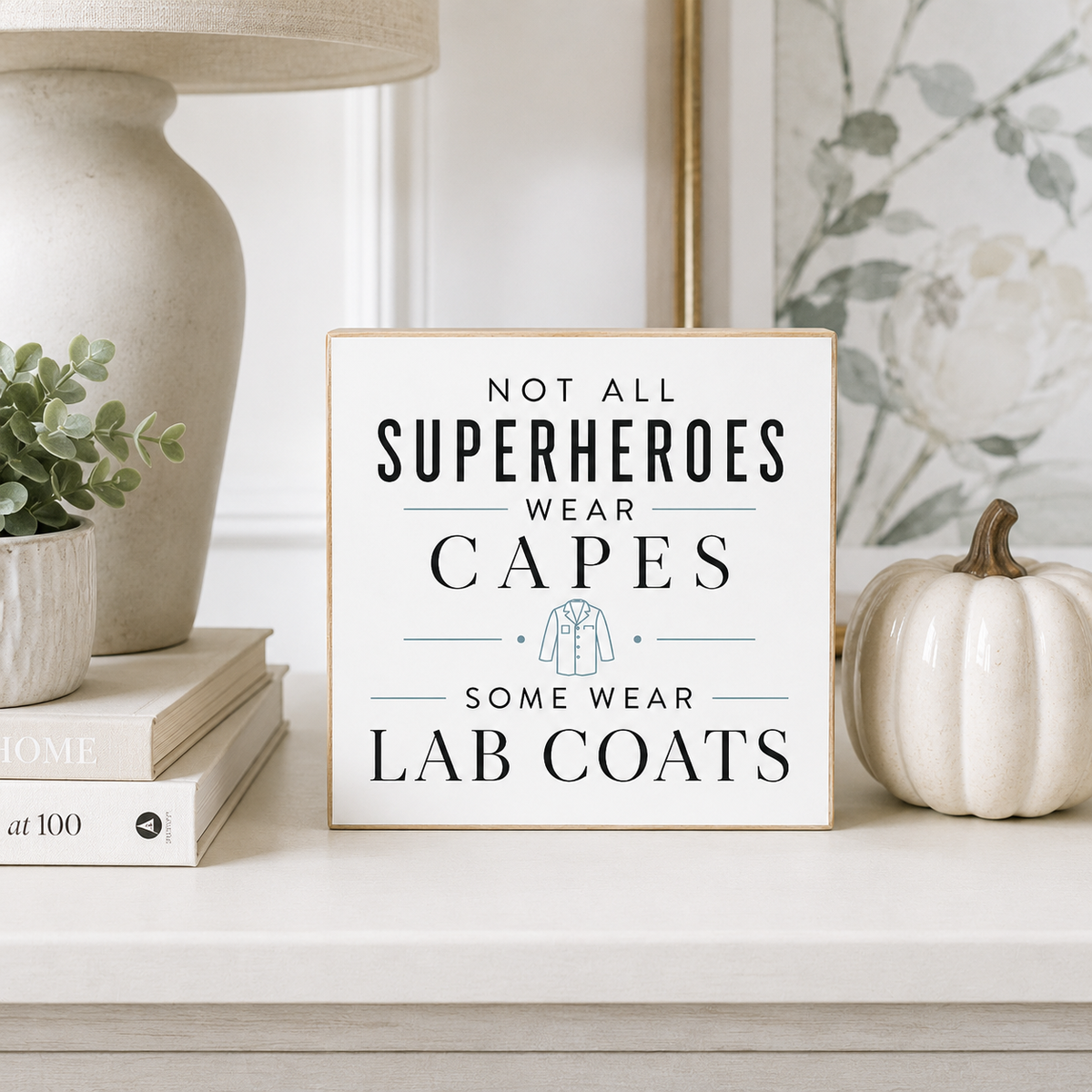 A square decorative sign with a light wood frame and white background reading 'NOT ALL SUPERHEROES WEAR CAPES SOME WEAR LAB COATS' in dark lettering, styled on a shelf beside a small white pumpkin, books, and a vase.