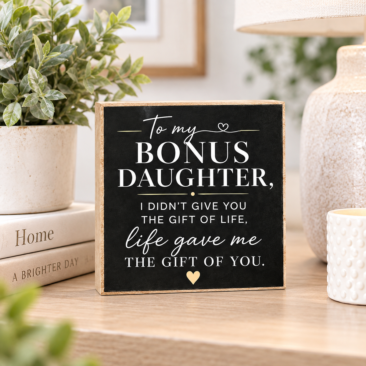 A square black decorative sign with white text reading, 'To my BONUS DAUGHTER, I DIDN'T GIVE YOU THE GIFT OF LIFE, life gave me THE GIFT OF YOU.' It is styled on a wooden table with a potted plant, books, a lamp, and a candle in a home setting.