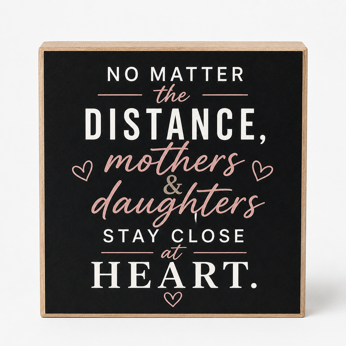 Square wooden sign with a black face and natural wood edges, featuring the phrase 'NO MATTER the DISTANCE, mothers & daughters STAY CLOSE at HEART.' in white and blush pink lettering with small heart icons, shown on a light gray background.