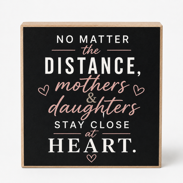 Square wooden sign with a black face and natural wood edges, featuring the phrase 'NO MATTER the DISTANCE, mothers & daughters STAY CLOSE at HEART.' in white and blush pink lettering with small heart icons, shown on a light gray background.
