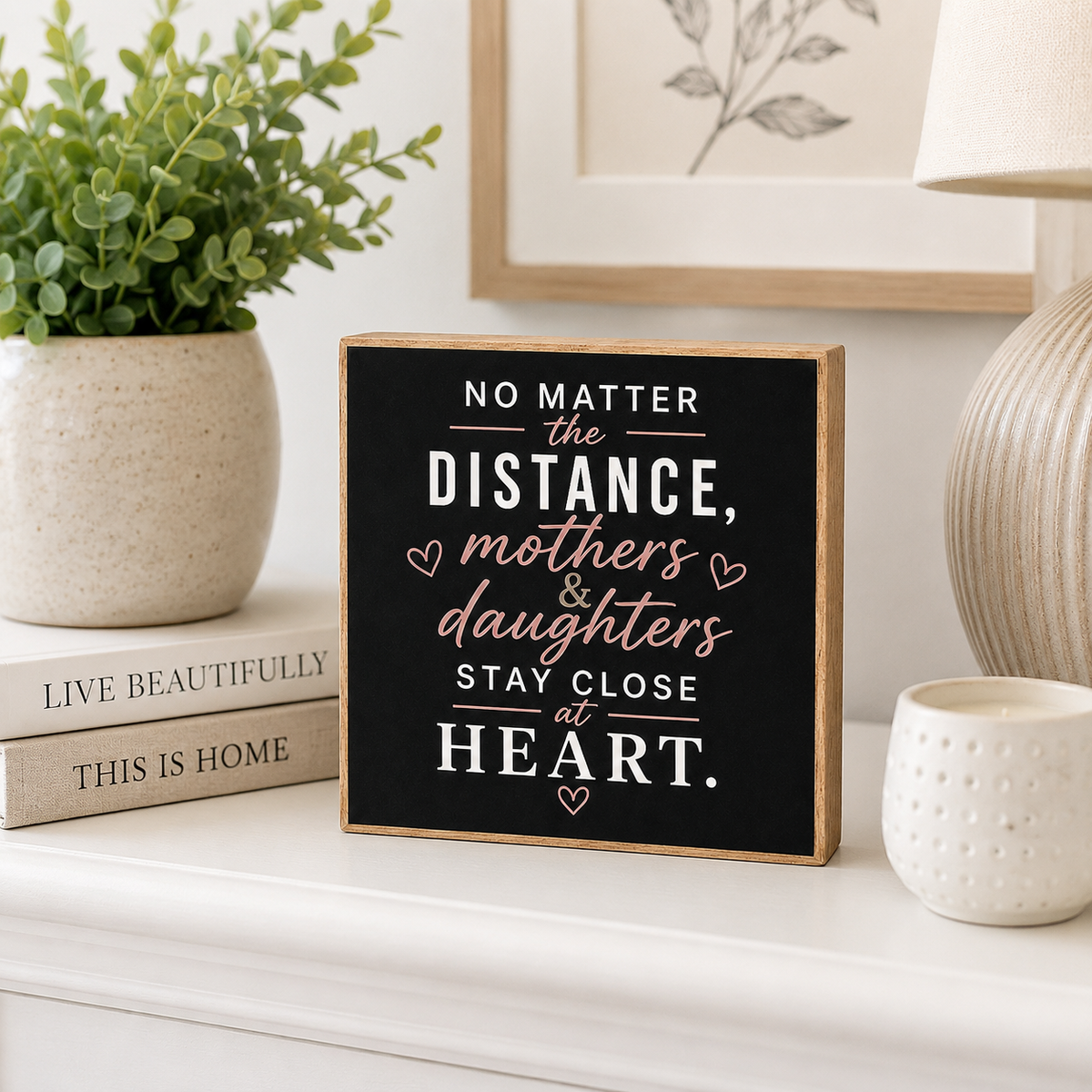 A square wooden sign with a black face and natural wood edges, featuring the quote 'NO MATTER the DISTANCE, mothers & daughters STAY CLOSE at HEART.' in white and blush pink lettering.