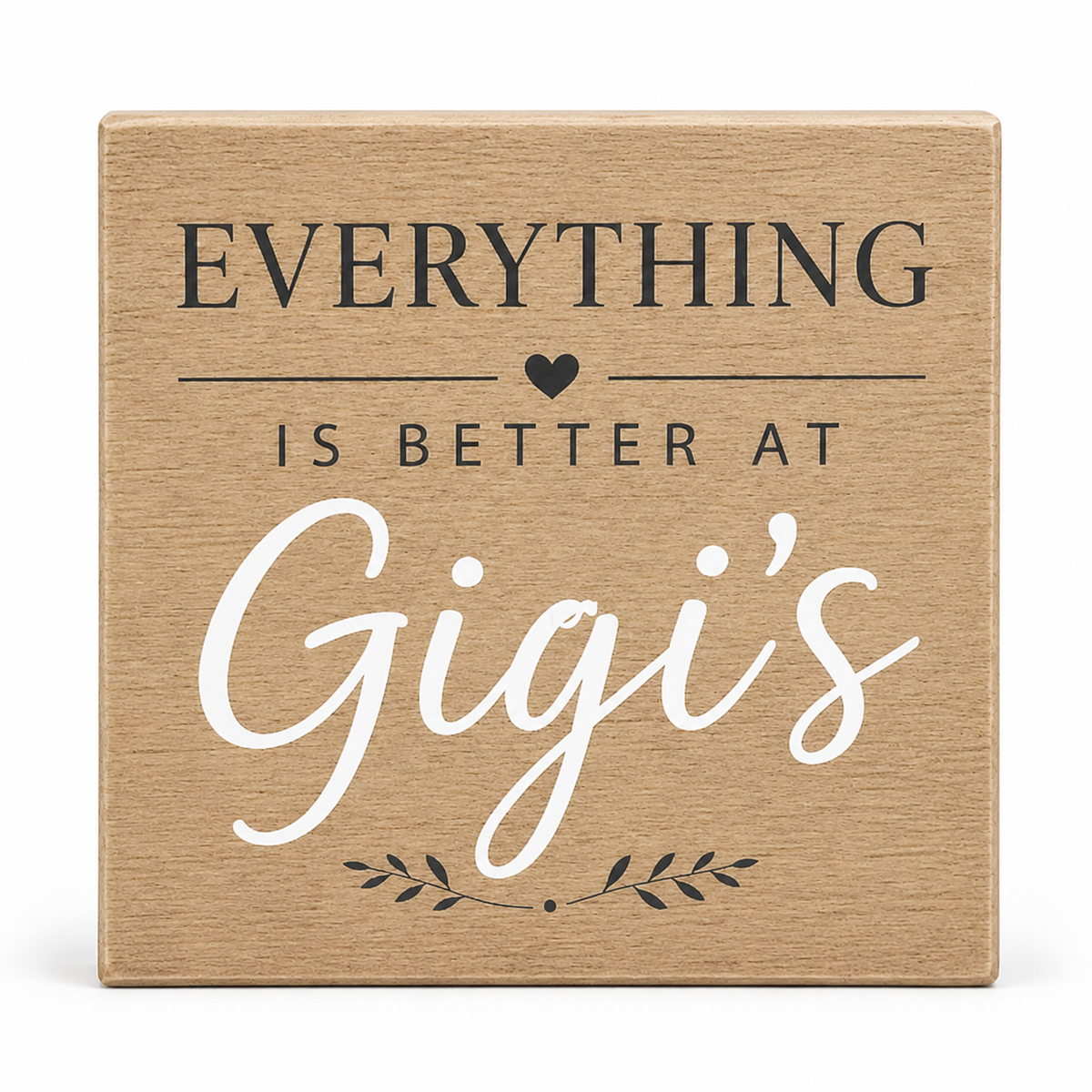 A square wooden sign with a natural light brown wood grain finish. It features the printed text 'EVERYTHING IS BETTER AT Gigi's' in dark gray and white lettering, with a small black heart and leafy accent.