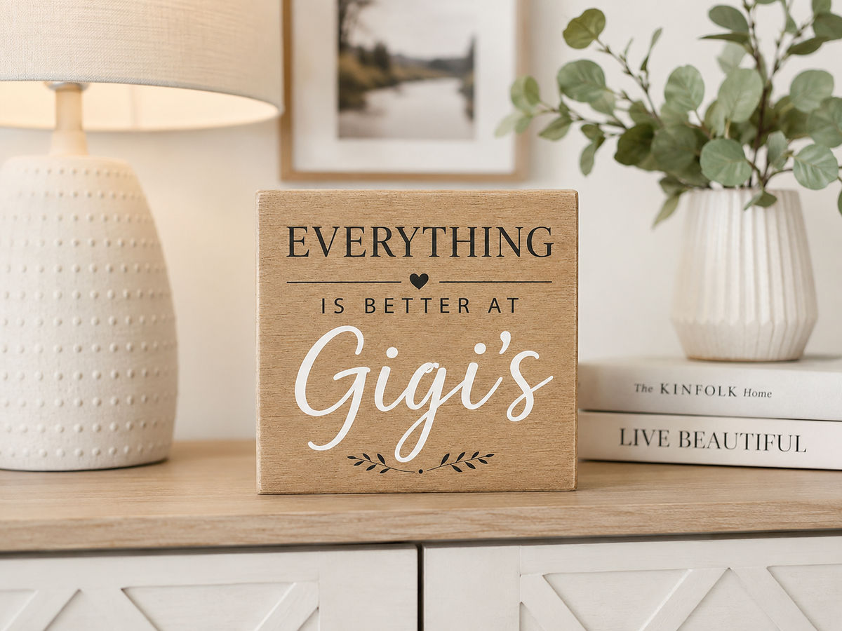 A square wooden sign with the text 'EVERYTHING IS BETTER AT Gigi's' in black and white lettering, styled on a light wood cabinet next to a lamp, books, and a vase of greenery.