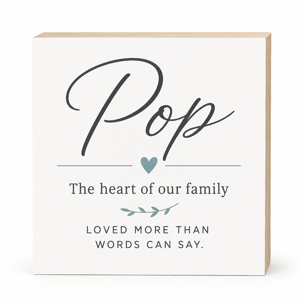 A square white decorative sign with a light wood edge, featuring the word 'Pop' in large black script, a small blue heart, and the phrases 'The heart of our family' and 'LOVED MORE THAN WORDS CAN SAY.' on a clean white background.