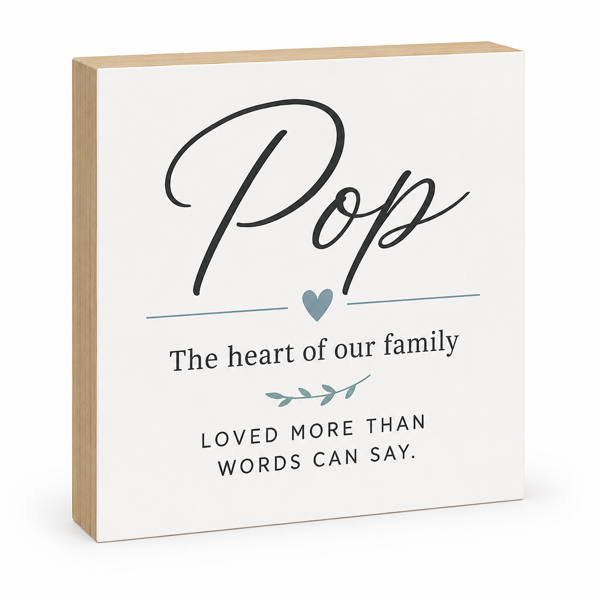 A square decorative wall sign with a white front panel and natural wood sides. The sign features the word 'Pop' in large black script, a small blue heart, and the phrases 'The heart of our family' and 'LOVED MORE THAN WORDS CAN SAY.