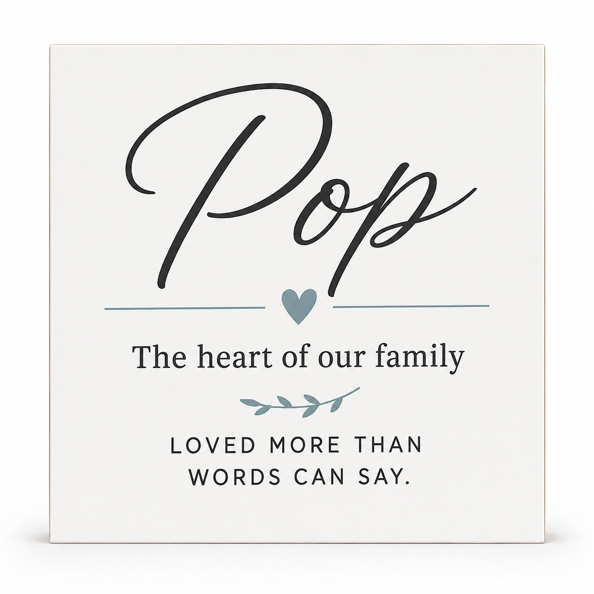 A square white decorative sign with printed text reading 'Pop' in large black script, followed by 'The heart of our family,' a small blue heart, a leaf motif, and the phrase 'LOVED MORE THAN WORDS CAN SAY.' on a clean white background.