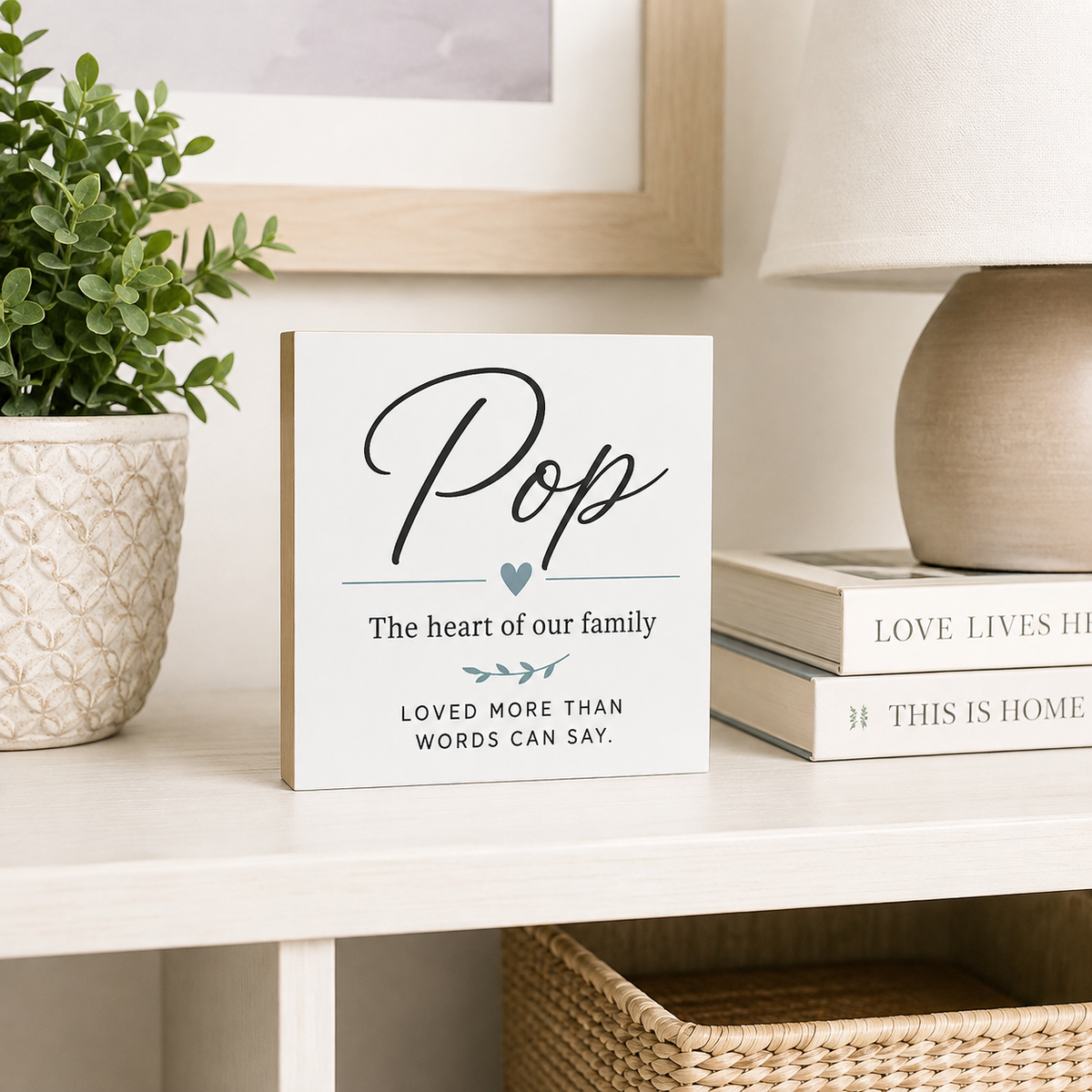 A square white decorative sign with black script reading 'Pop' and smaller family-themed text, styled on a light wood shelf beside a potted plant, books, and a lamp.