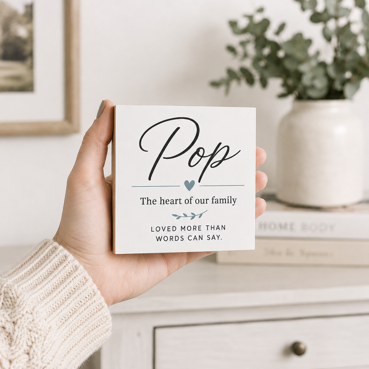 A hand wearing a cream knit sweater holds a square white gift box with the word 'Pop' in large script and the phrase 'The heart of our family' on the front. The background shows a softly blurred home interior with a plant in a white vase and a light-colored cabinet.