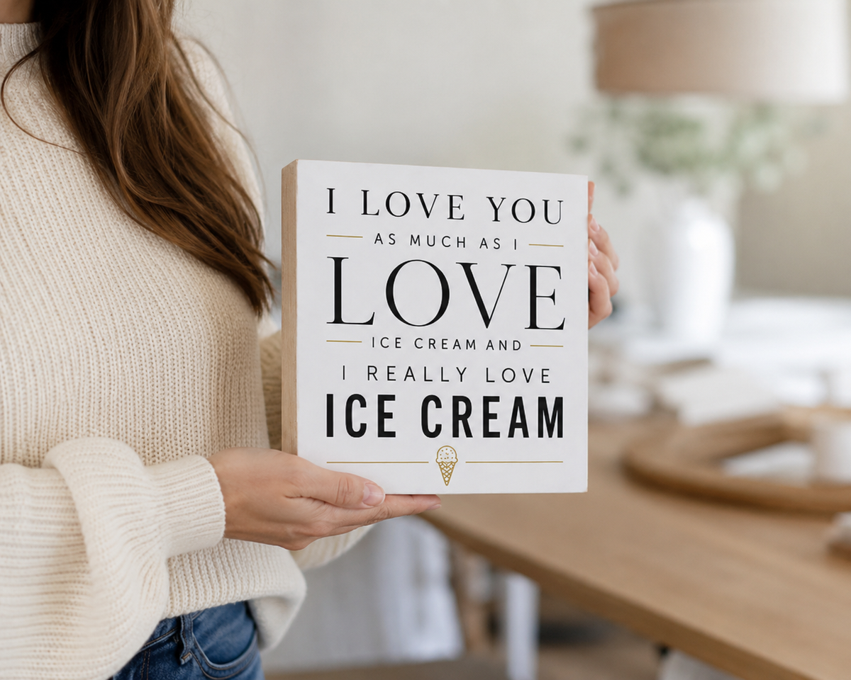 A person in a cream knit sweater holds a square white decorative sign with black text that reads, 'I LOVE YOU AS MUCH AS I LOVE ICE CREAM AND I REALLY LOVE ICE CREAM,' with a small ice cream cone graphic at the bottom. The background is a softly blurred indoor room with a wooden table and lamp.