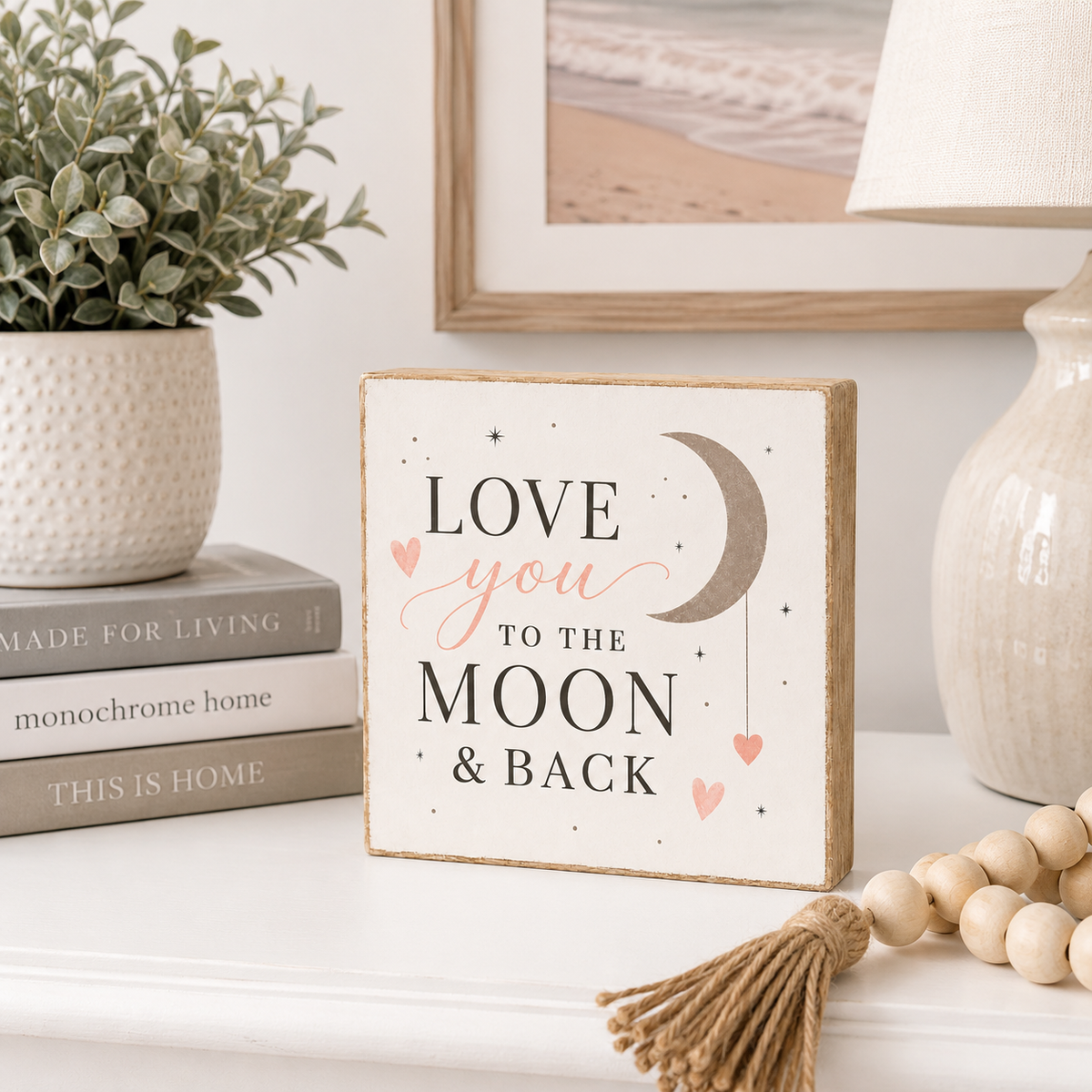 A square decorative wooden sign with a natural jute border and the phrase 'LOVE you TO THE MOON & BACK' in black and pink lettering, styled on a white shelf with a plant, books, a lamp, and wooden beads.