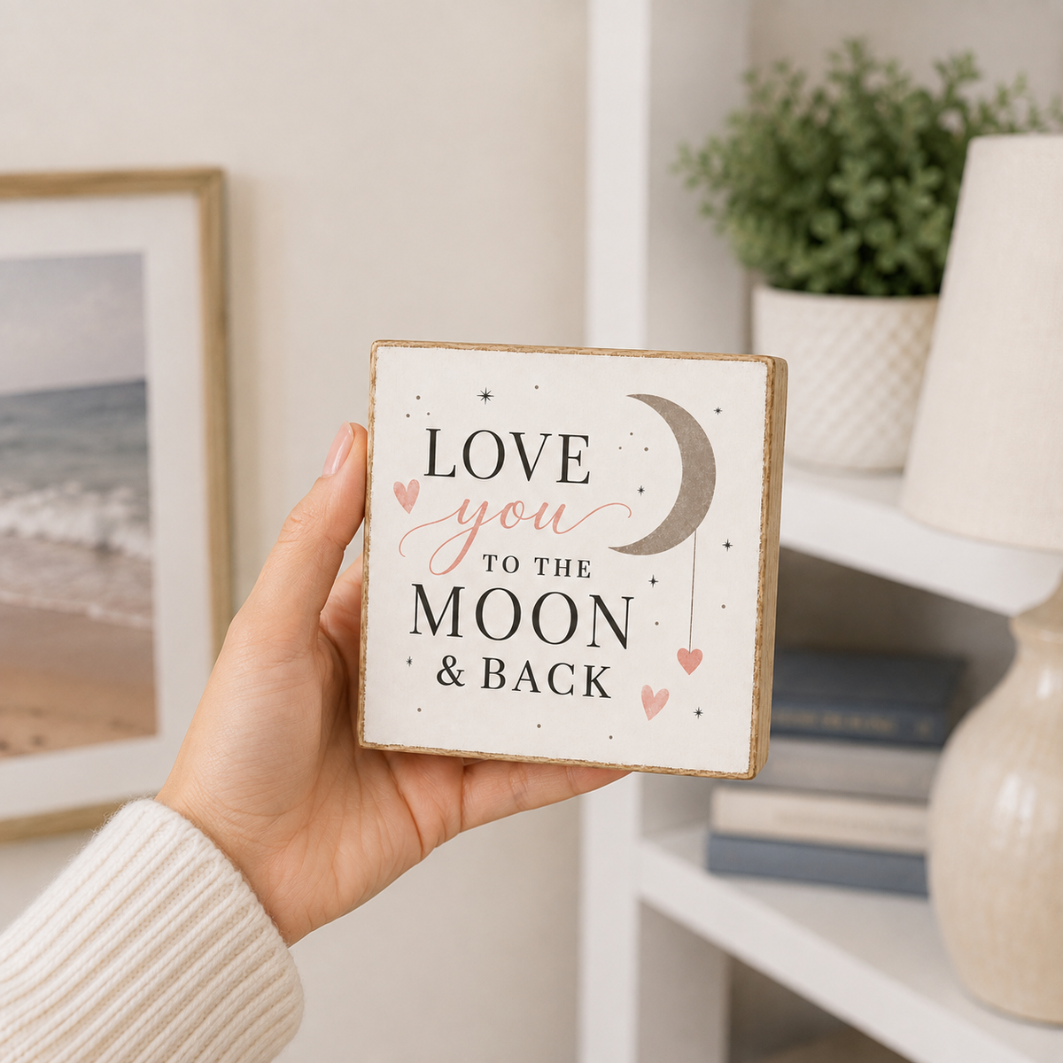 A hand holds a square decorative wooden sign that reads 'LOVE you TO THE MOON & BACK' with a crescent moon and small hearts. The sign is shown in a softly lit room with shelves, books, and a plant in the background.