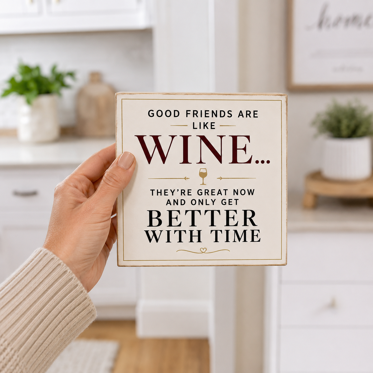 A hand wearing a beige ribbed sweater holds a square decorative sign with a rustic cream background and dark red and black text that reads, 'GOOD FRIENDS ARE LIKE WINE.