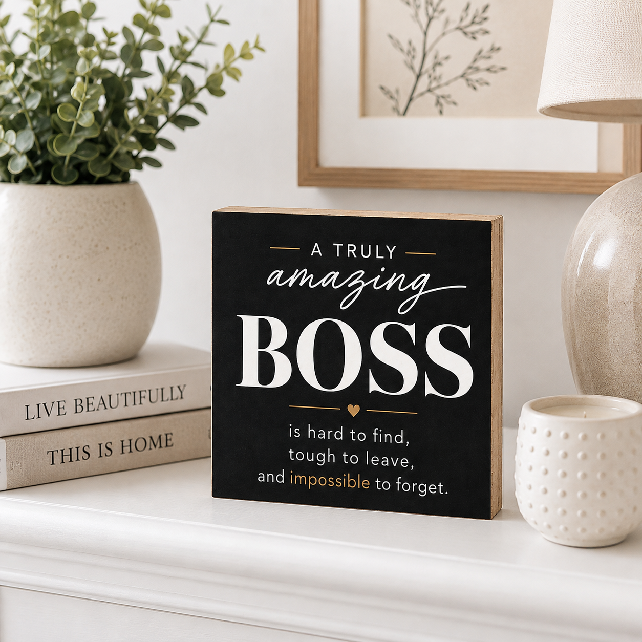 A rectangular black decorative sign with a light wood border and white text reading “A TRULY amazing BOSS is hard to find, tough to leave, and impossible to forget.” It is styled on a white mantel with a potted plant, books, a candle, and a lamp in a home interior setting.