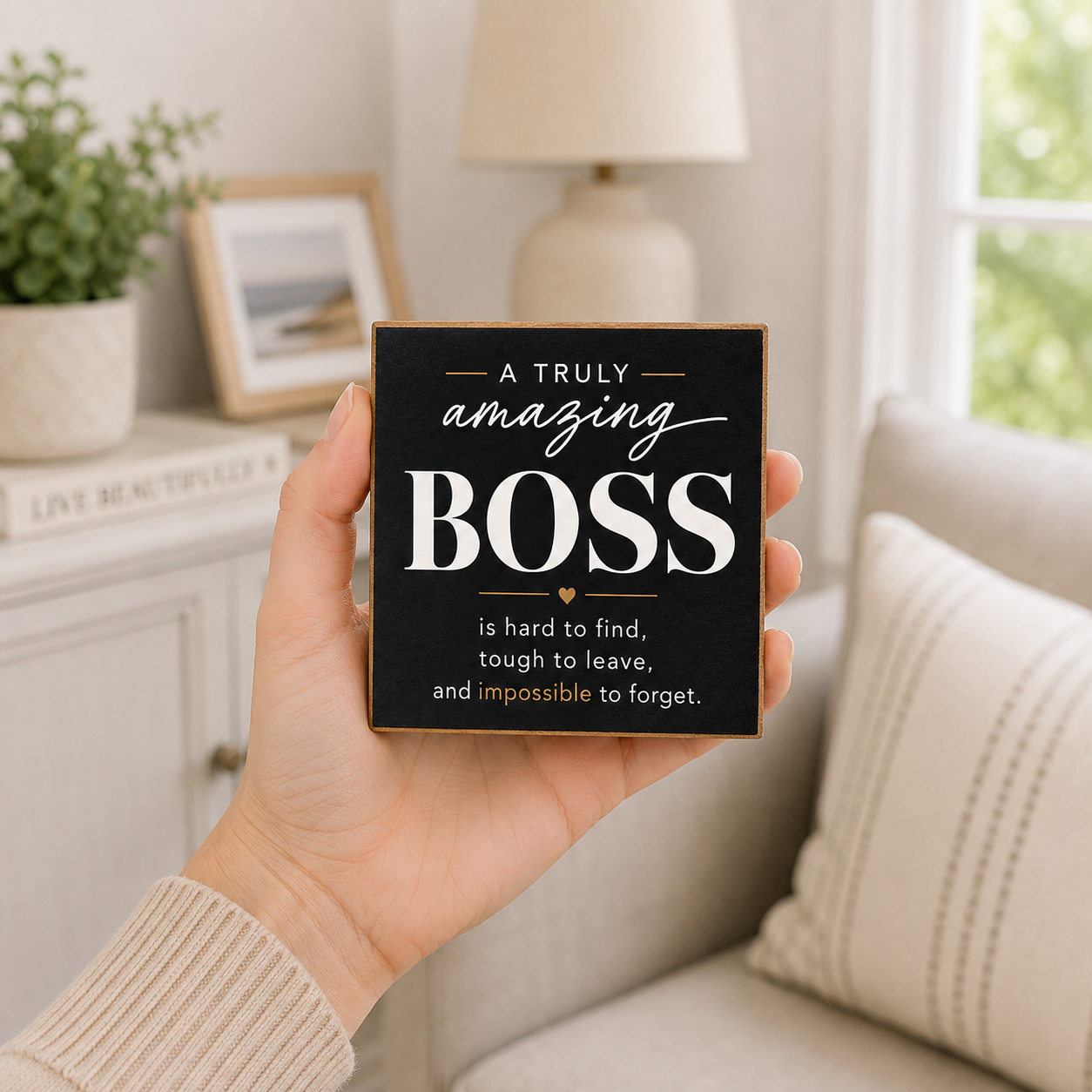 A hand holds a square black decorative sign with a gold border and white text that reads, 'A TRULY amazing BOSS is hard to find, tough to leave, and impossible to forget.' The background shows a softly lit living room with a sofa, lamp, framed picture, and plant.