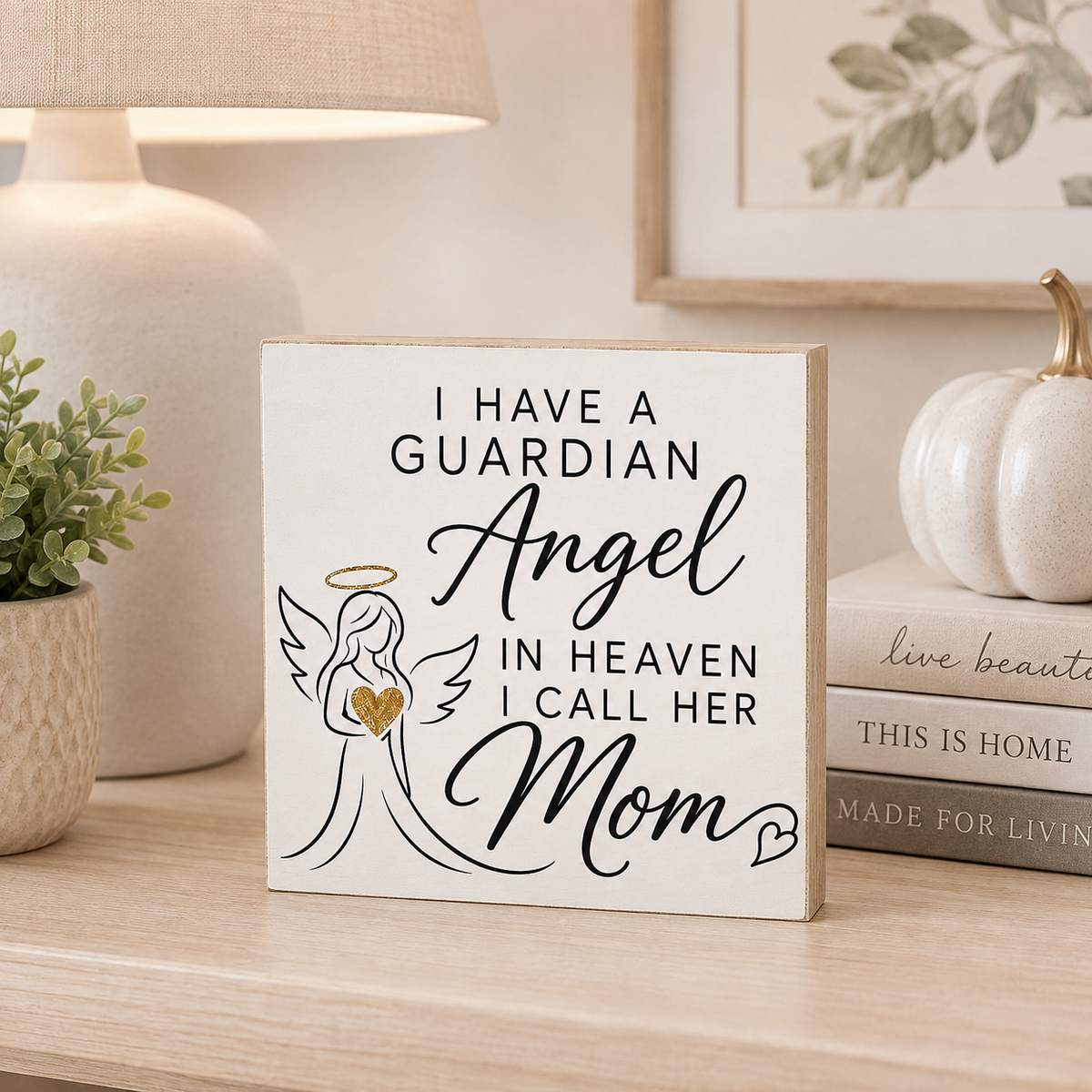 A square decorative wall sign with a cream background and natural wood edges, printed with the phrase 'I HAVE A GUARDIAN Angel IN HEAVEN I CALL HER Mom' in black script and serif text, plus a simple angel illustration holding a gold heart. The sign is styled on a wooden shelf beside a lamp, framed art, a small plant, and stacked books.